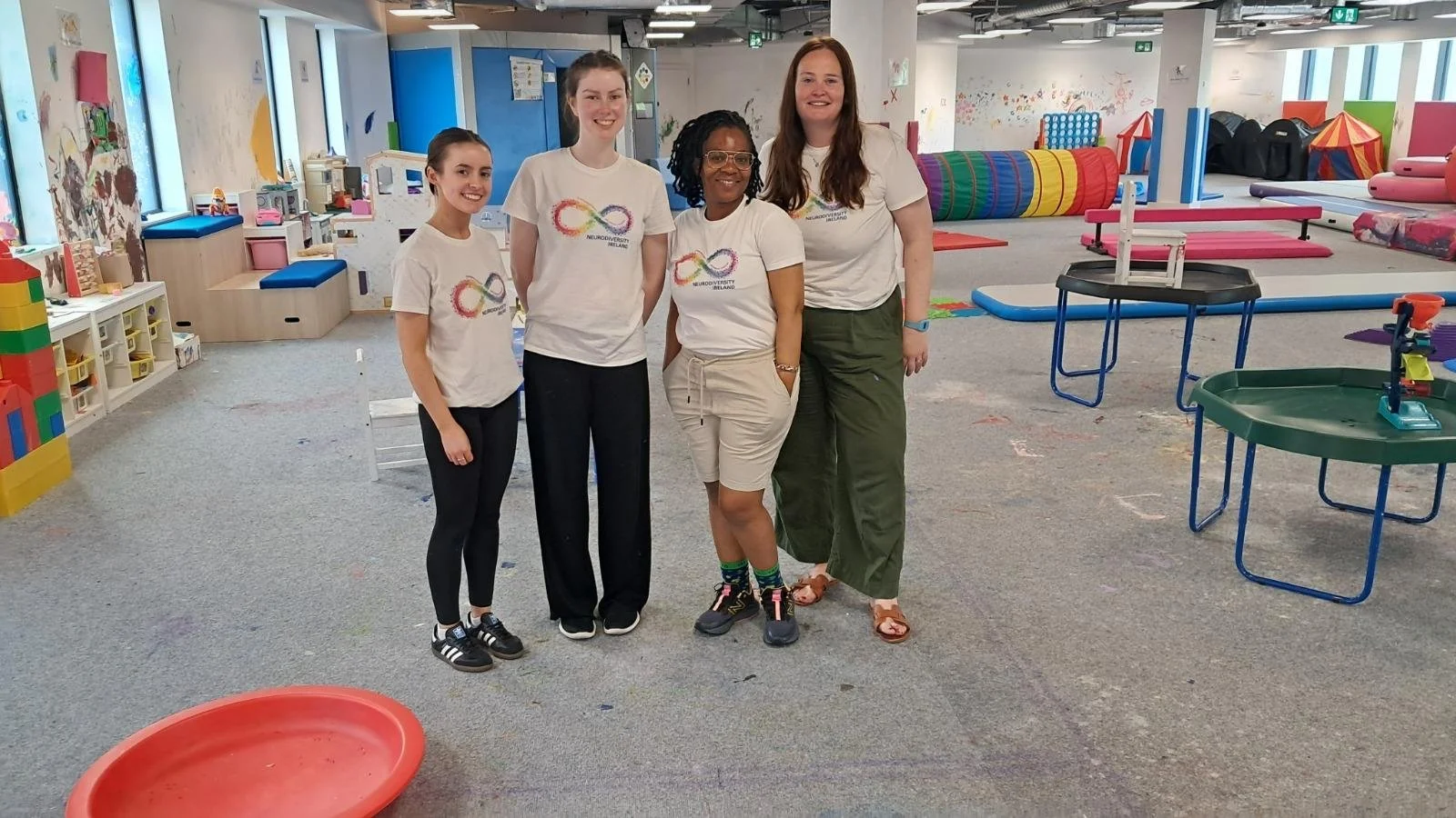 Four women standing together inside a colorful, child-friendly indoor playground or therapy center, smiling at the camera, wearing white t-shirts with a rainbow-colored infinity logo.