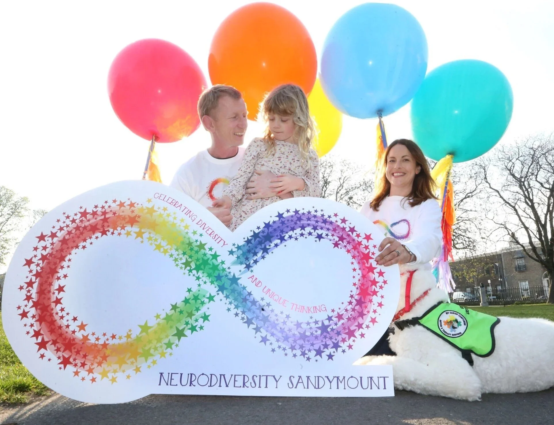 People holding a rainbow-colored sign with an infinity symbol and text about celebrating diversity and unique thinking, with balloons and a dog wearing a vest, outdoors during daytime.
