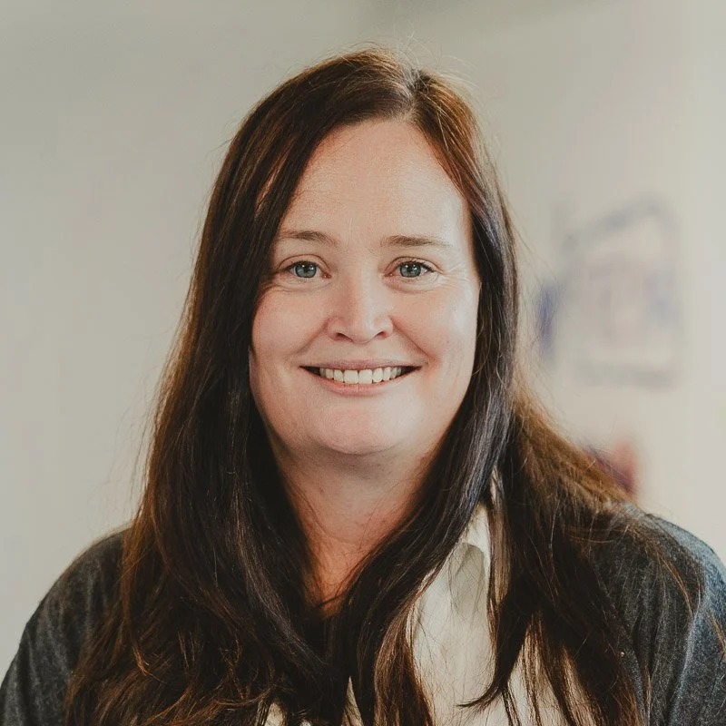A woman with long brown hair and blue eyes smiling at the camera, wearing a white blouse and a dark cardigan, in an indoor setting.