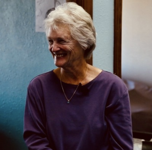 A smiling elderly woman with short, white hair wearing a purple top and a gold necklace, indoors with a blue wall and a mirror behind her.