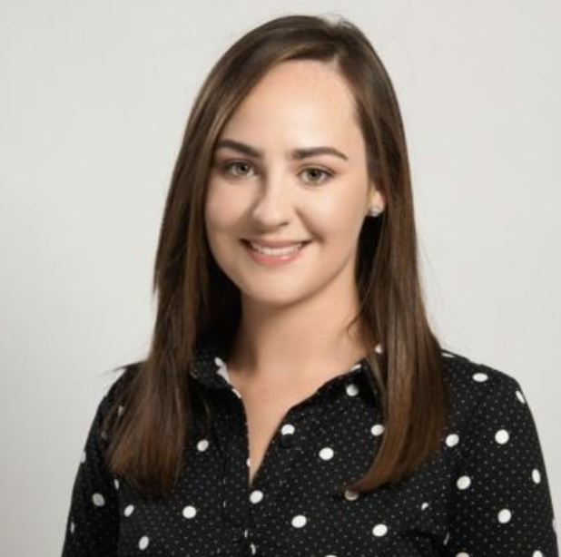 Portrait of a young woman with long brown hair, wearing a black blouse with white polka dots, smiling against a plain light background.