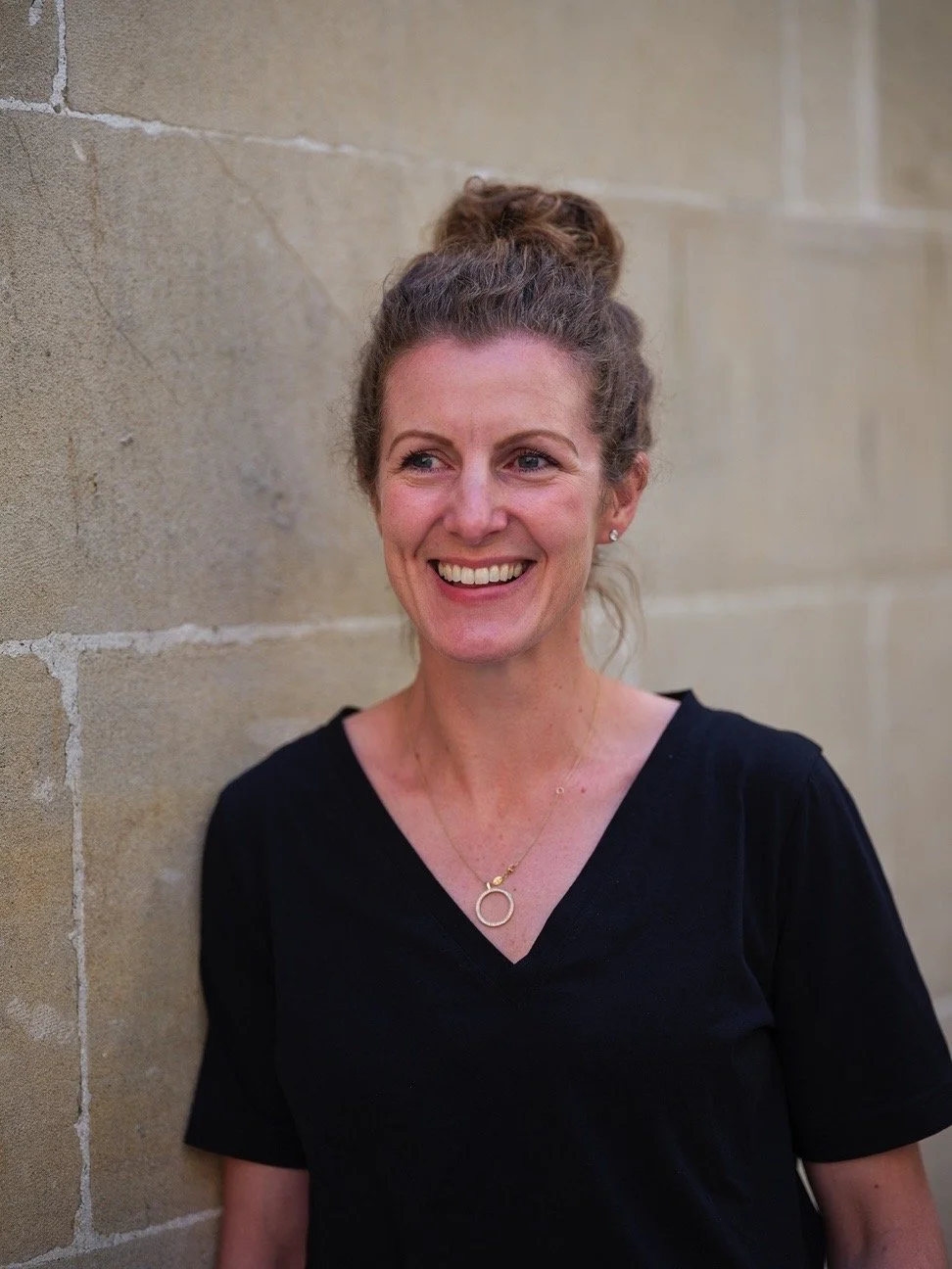 A smiling woman with brown hair in a bun, wearing a black shirt and a gold necklace, standing against a beige brick wall.