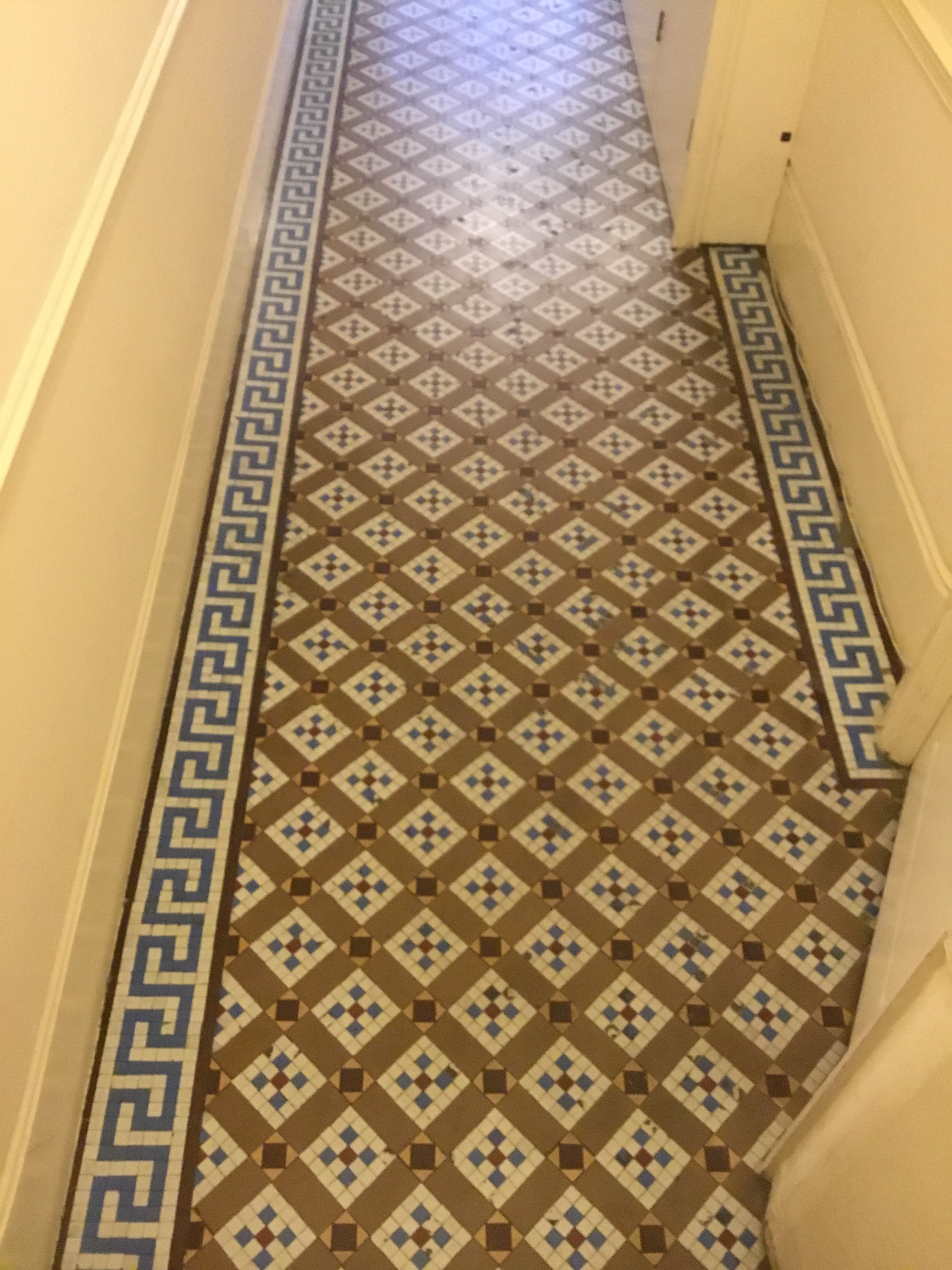 A patterned hallway floor with geometric tile designs in beige, brown, white, and blue, bordered by a Greek key pattern in blue and white.