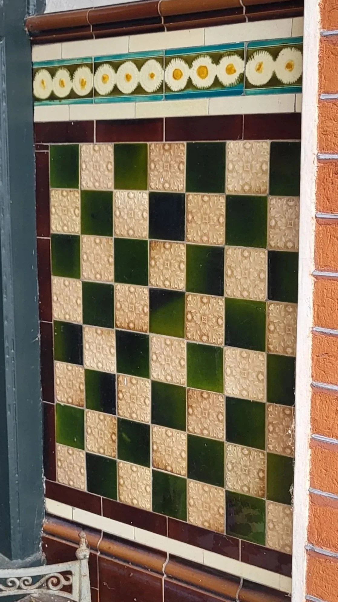Decorative Victorian tiled porch wall with a border of daisy flowers at the top, and a checkerboard pattern of beige and green tiles.