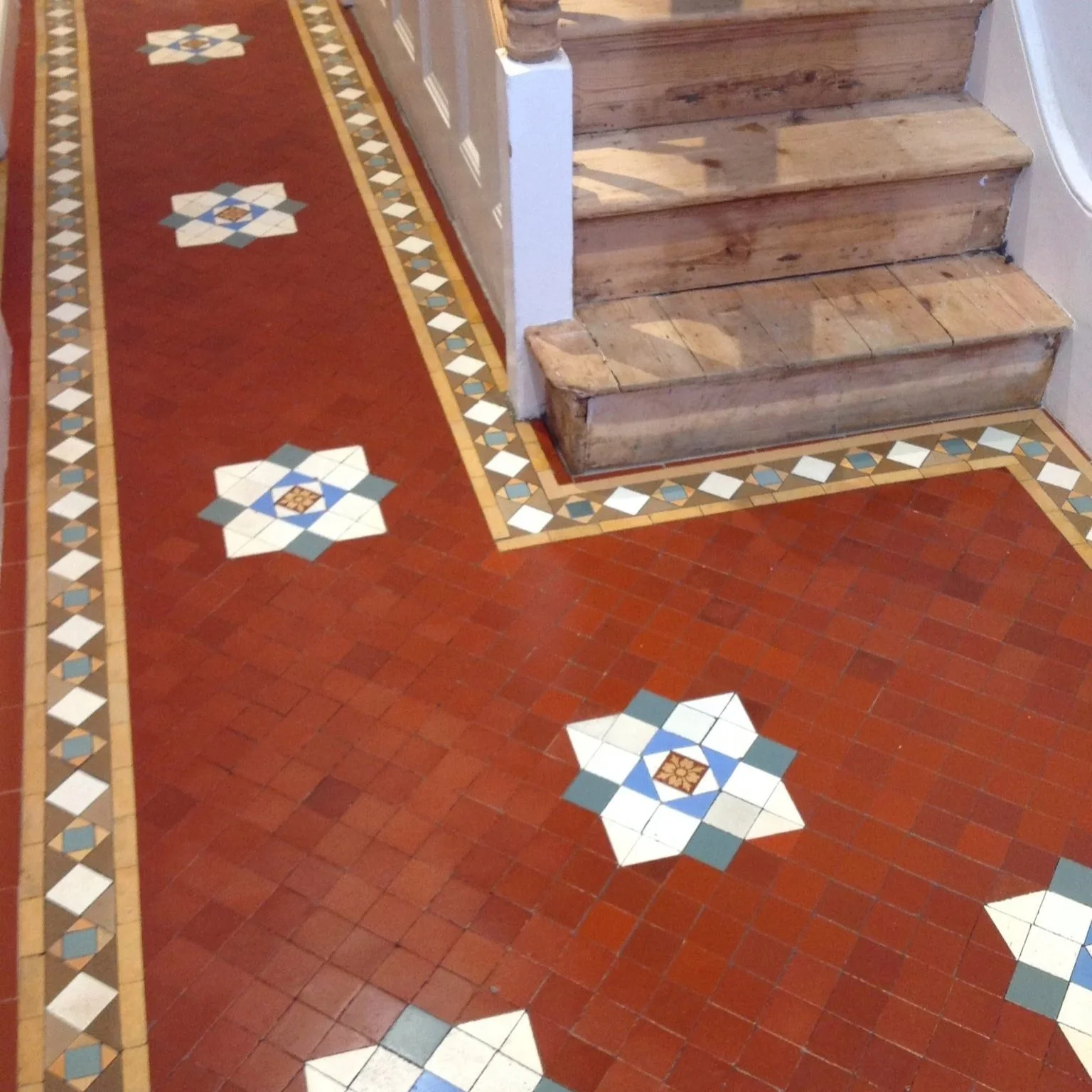 Decorative tile floor with red, beige, and blue geometric patterns, adjacent to wooden stairs in an interior space.