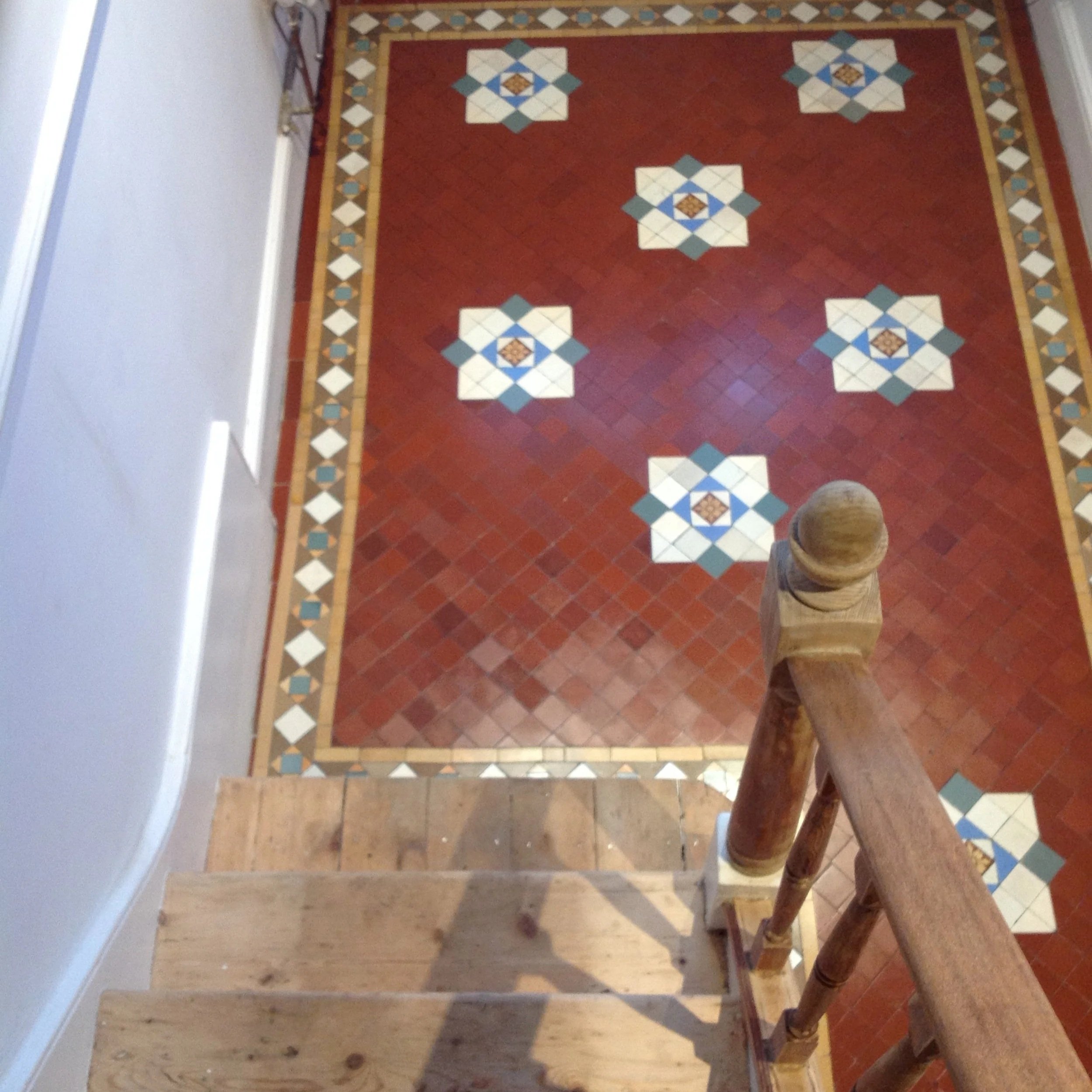 View of a staircase leading down to a patterned tile floor with a red background, featuring white, blue, and brown decorative motifs.