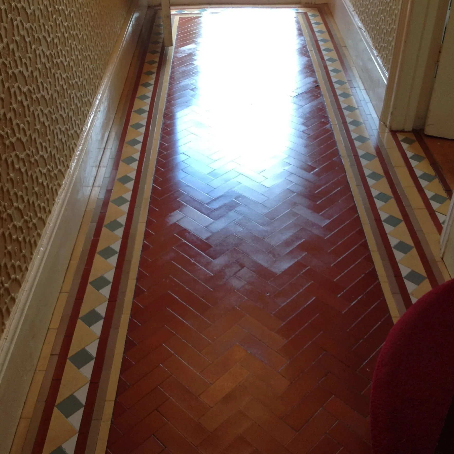 Glossy wooden herringbone-patterned floor with a decorative tile border along the edges, illuminated by sunlight from an open doorway.