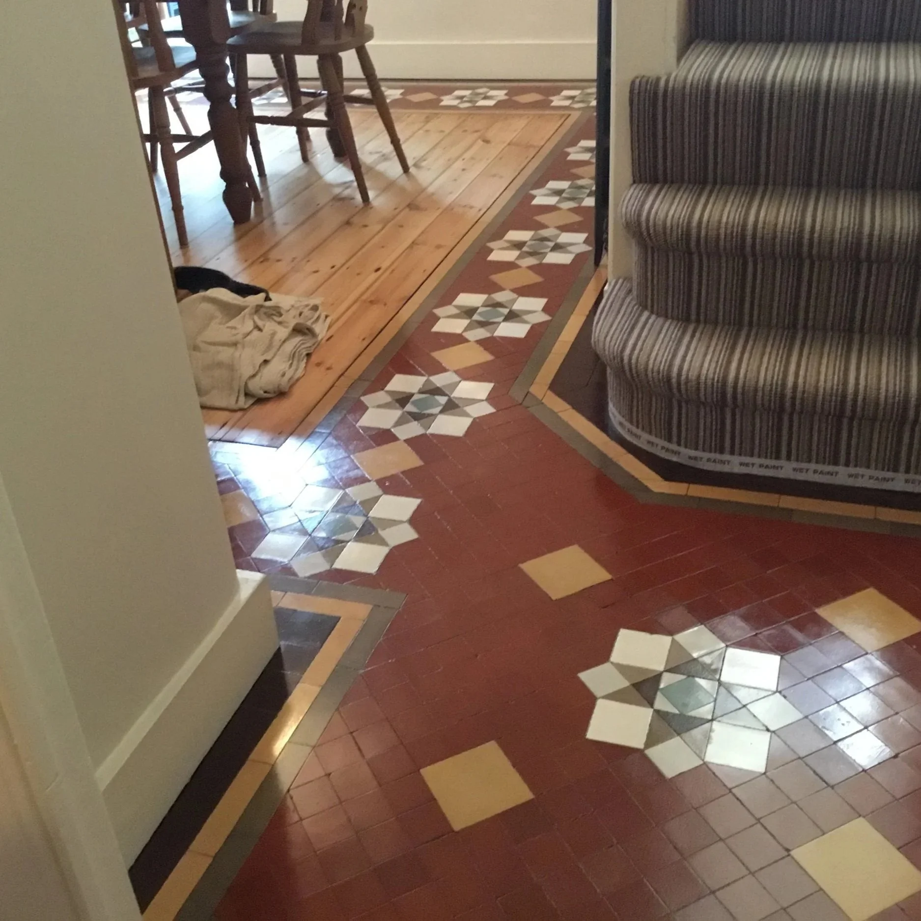 Interior image showing a floor with decorative tile pattern, a corner of a striped curved staircase on the right, and a glimpse of a dining area with wooden chairs in the background. There is a pile of clothes on the wooden floor near the wall on the