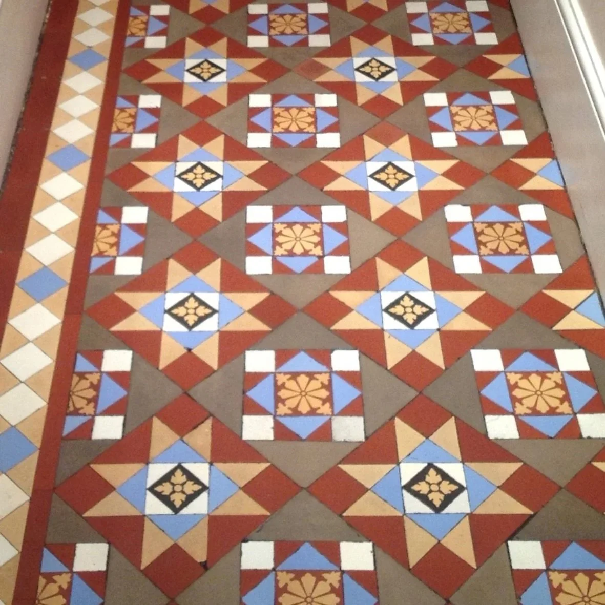 Colorful patterned tile floor with geometric designs in shades of red, blue, beige, black, and white, bordered by a strip of smaller square tiles.