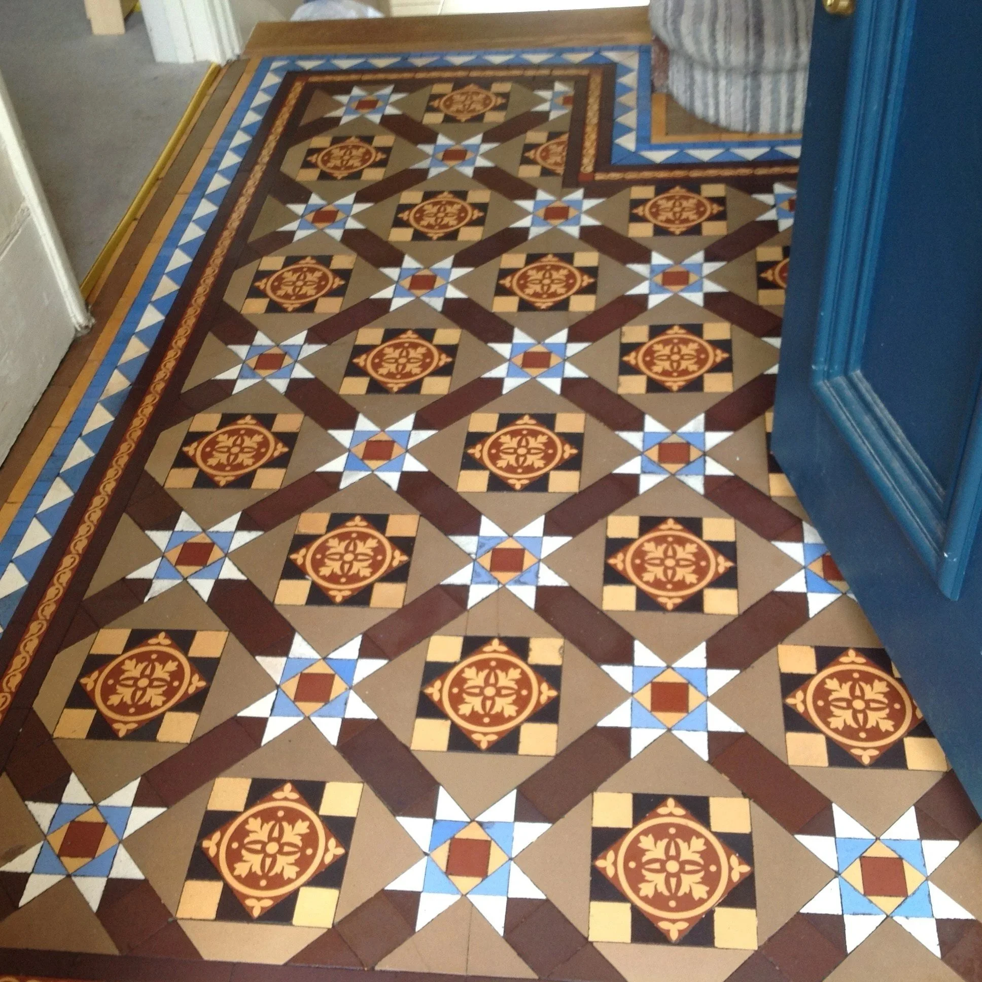 Patterned tile floor with geometric designs and border near a blue wall, partially visible door, and a striped object.