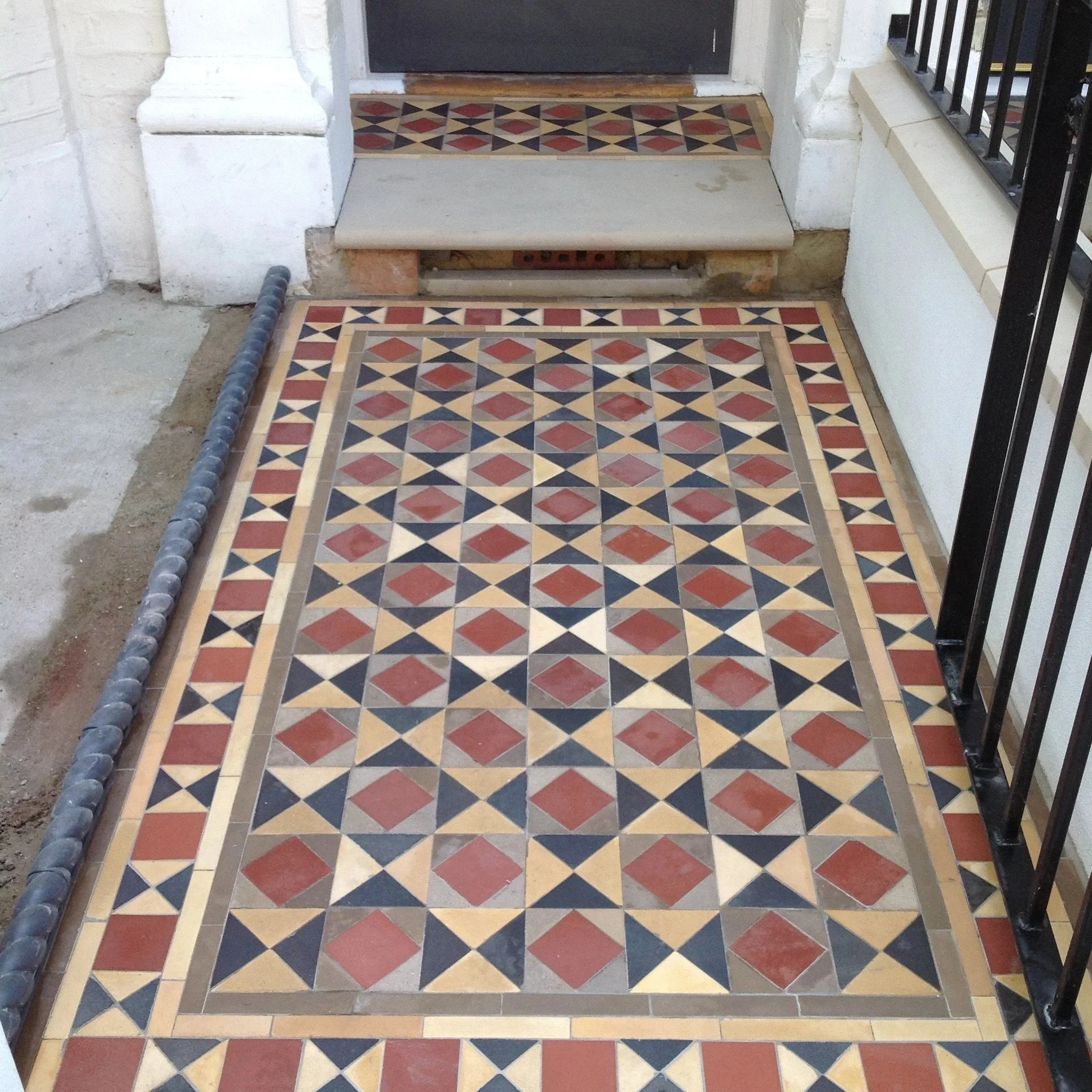 Colorful geometric tiled porch mat outside a doorway with stairs and a black metal railing.