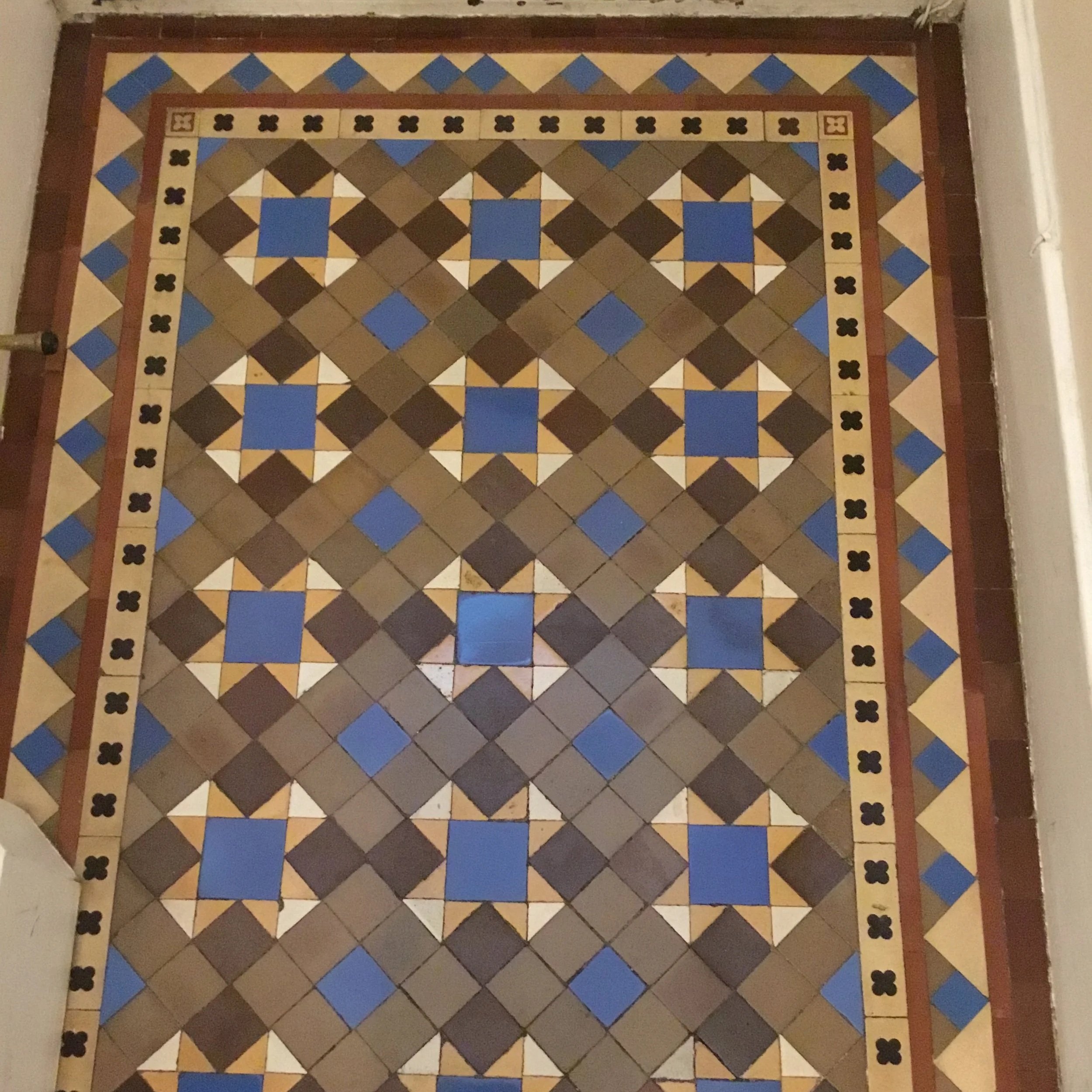 Decorative tile floor with geometric pattern in brown, blue, beige, and white colors, featuring a border with a floral motif.