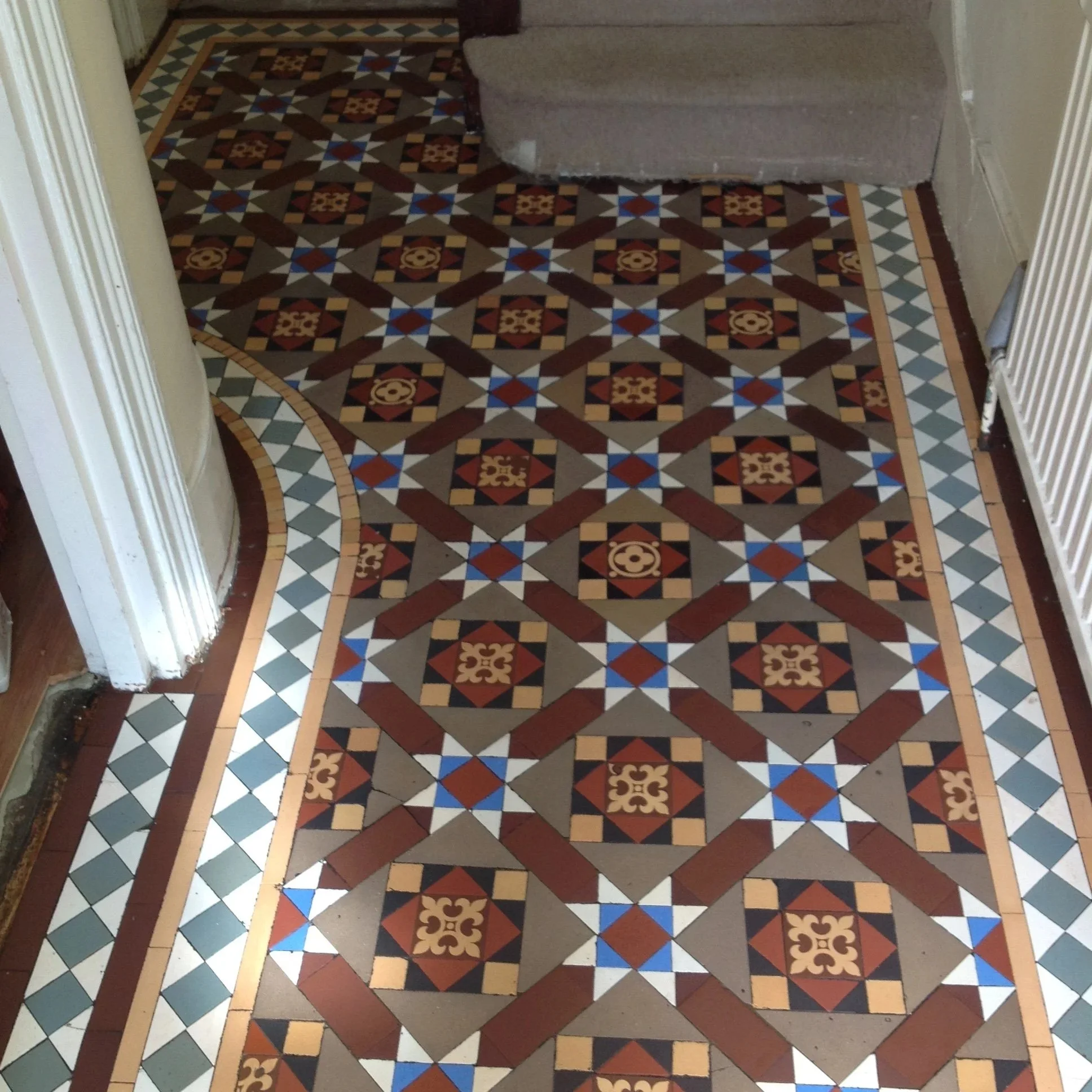 Decorative tiled floor with geometric patterns and intricate designs, bordered by a plain tile edge, in a hallway next to a staircase and a radiator.