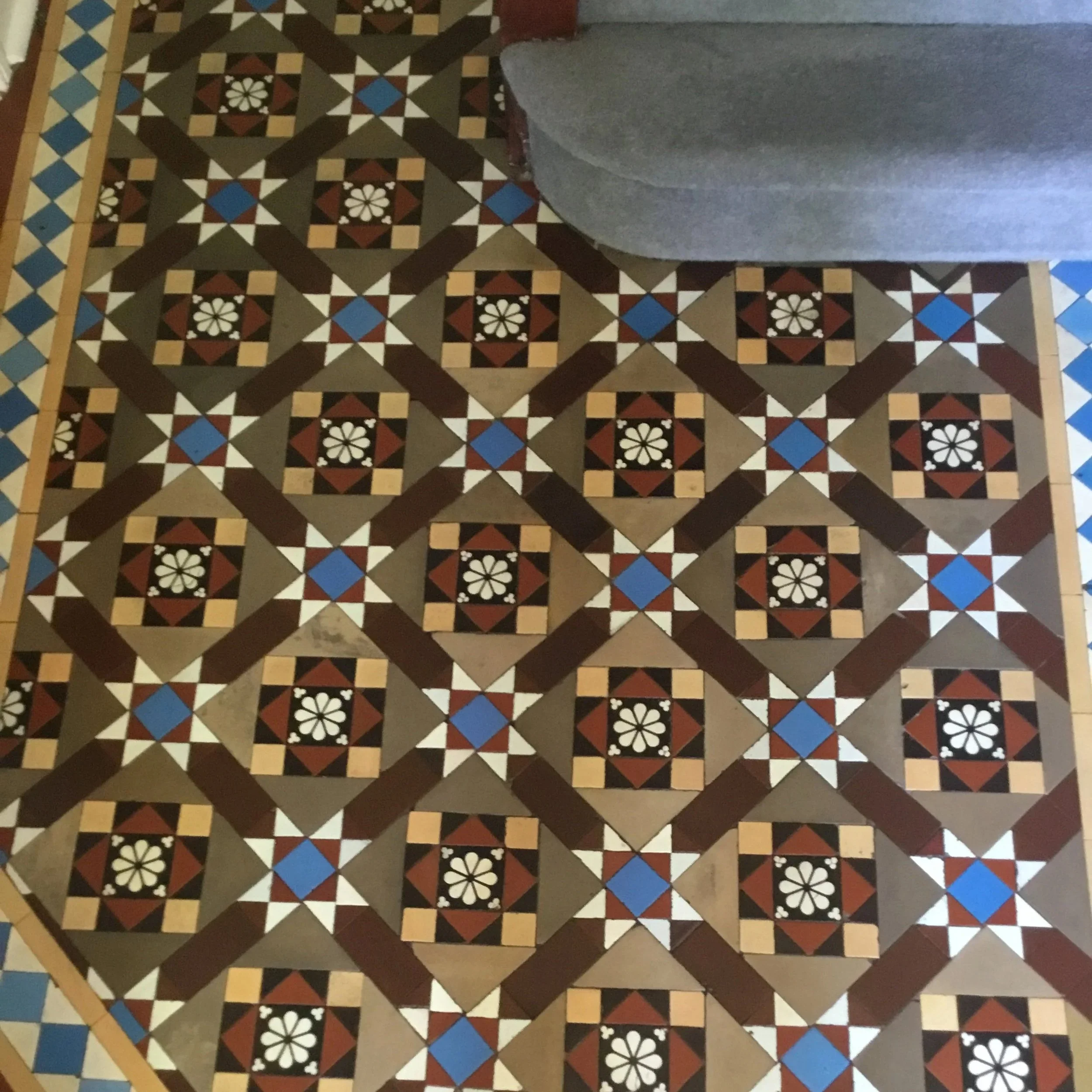 Patterned tile floor with geometric shapes in black, brown, beige, white, and blue, adjacent to a gray couch.