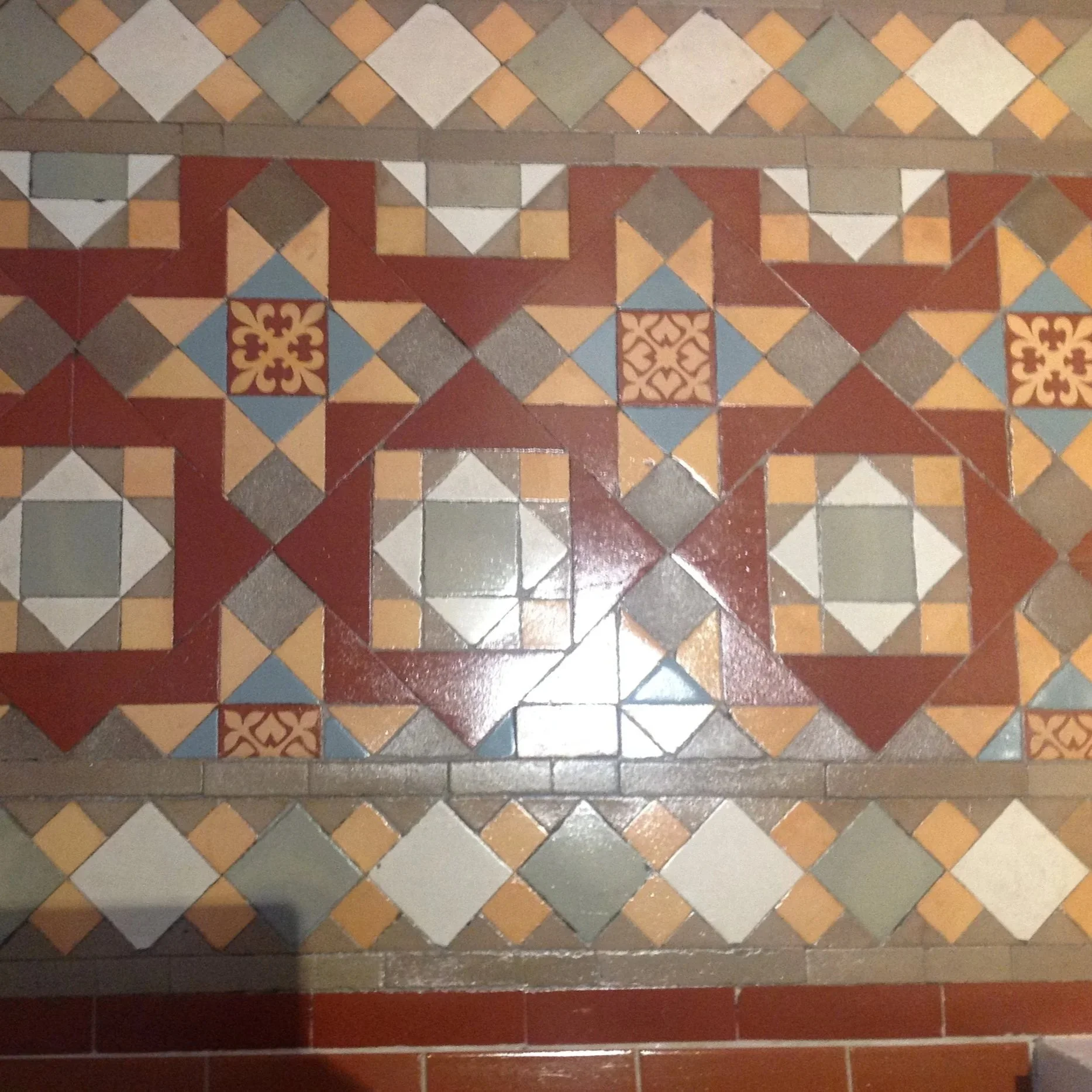 Patterned ceramic tiles forming intricate geometric and floral designs in shades of red, beige, gray, and blue, arranged in a symmetrical layout on a floor.