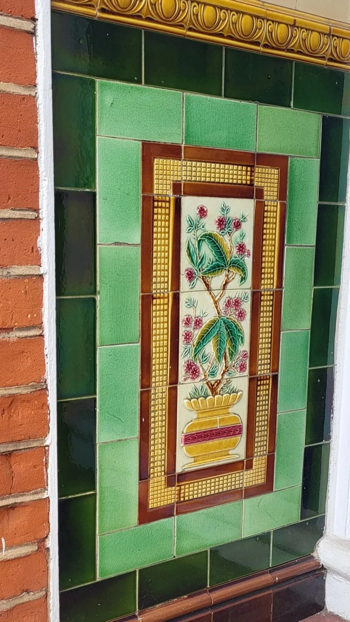 Decorative tile artwork of a potted plant with pink flowers and green leaves, framed by green and yellow tiles.