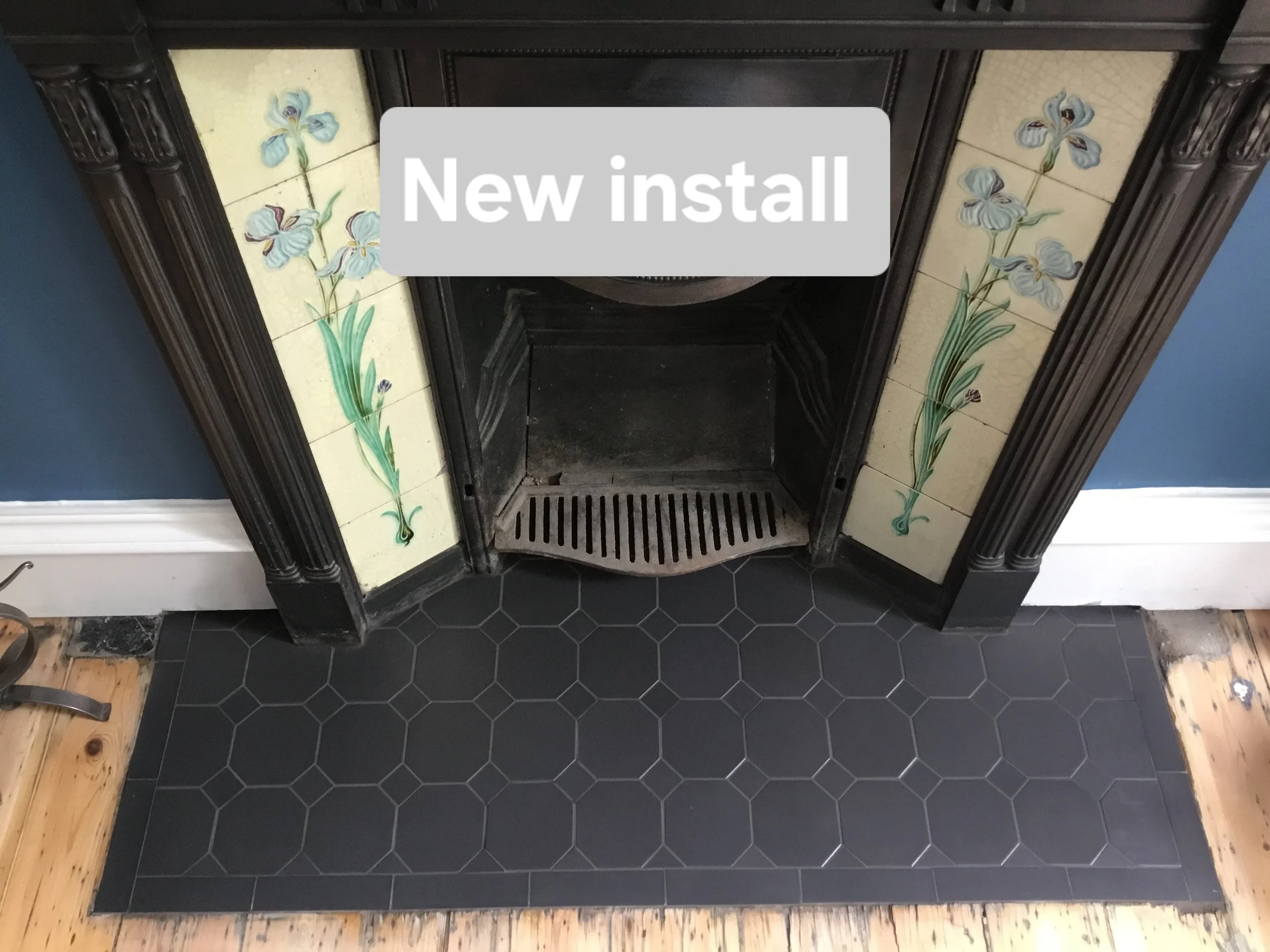 Fireplace with decorative tiles featuring blue iris flowers, a black surround, and black hexagonal tile hearth on a wooden floor, with a 'New install' label overlay.