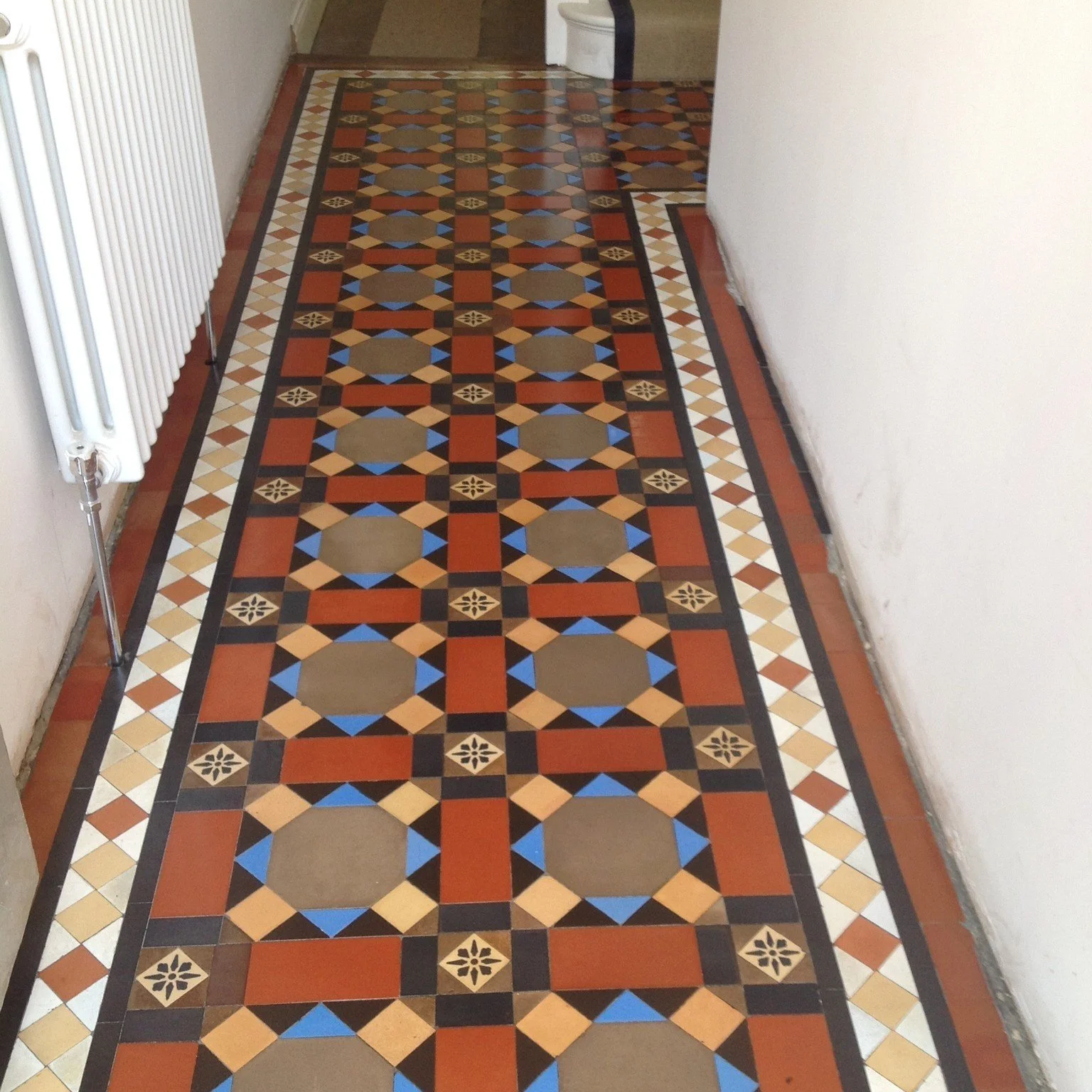 Colorful patterned tile floor in a hallway with a radiator on the left and a toilet in the background.
