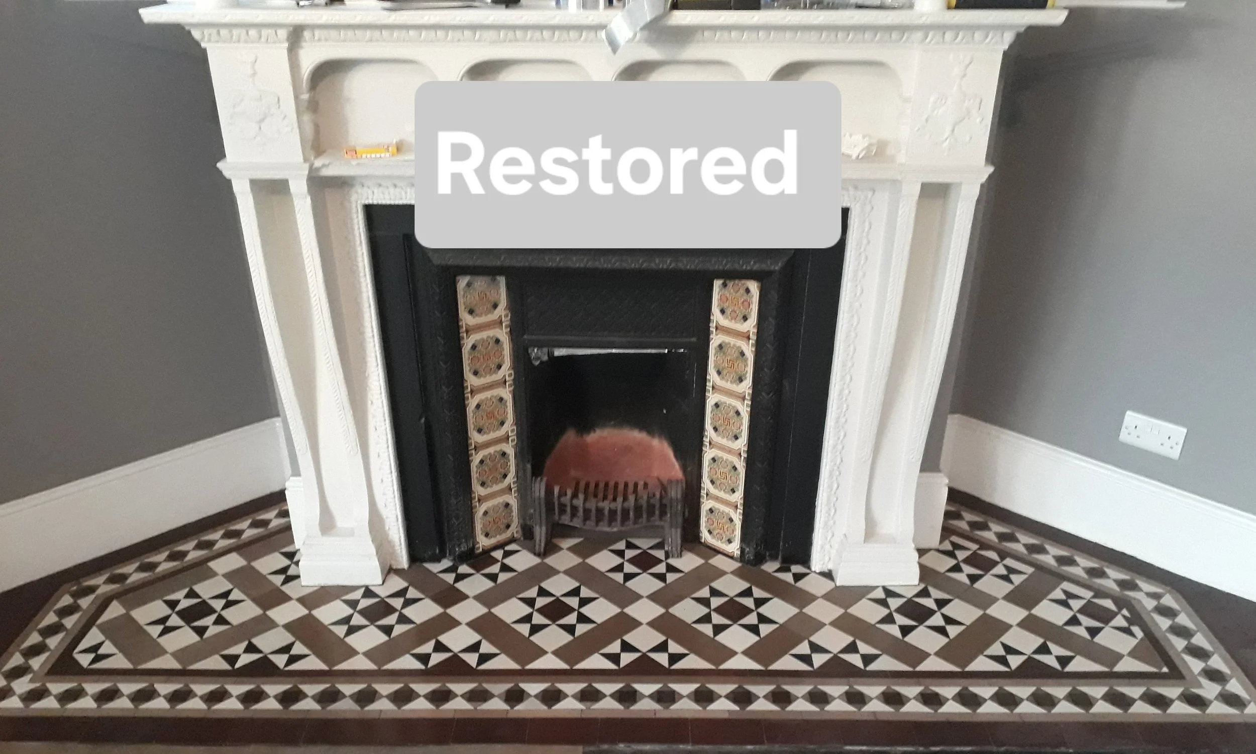 Restored fireplace with decorative tile border, white mantel, and patterned tile floor in a living room.