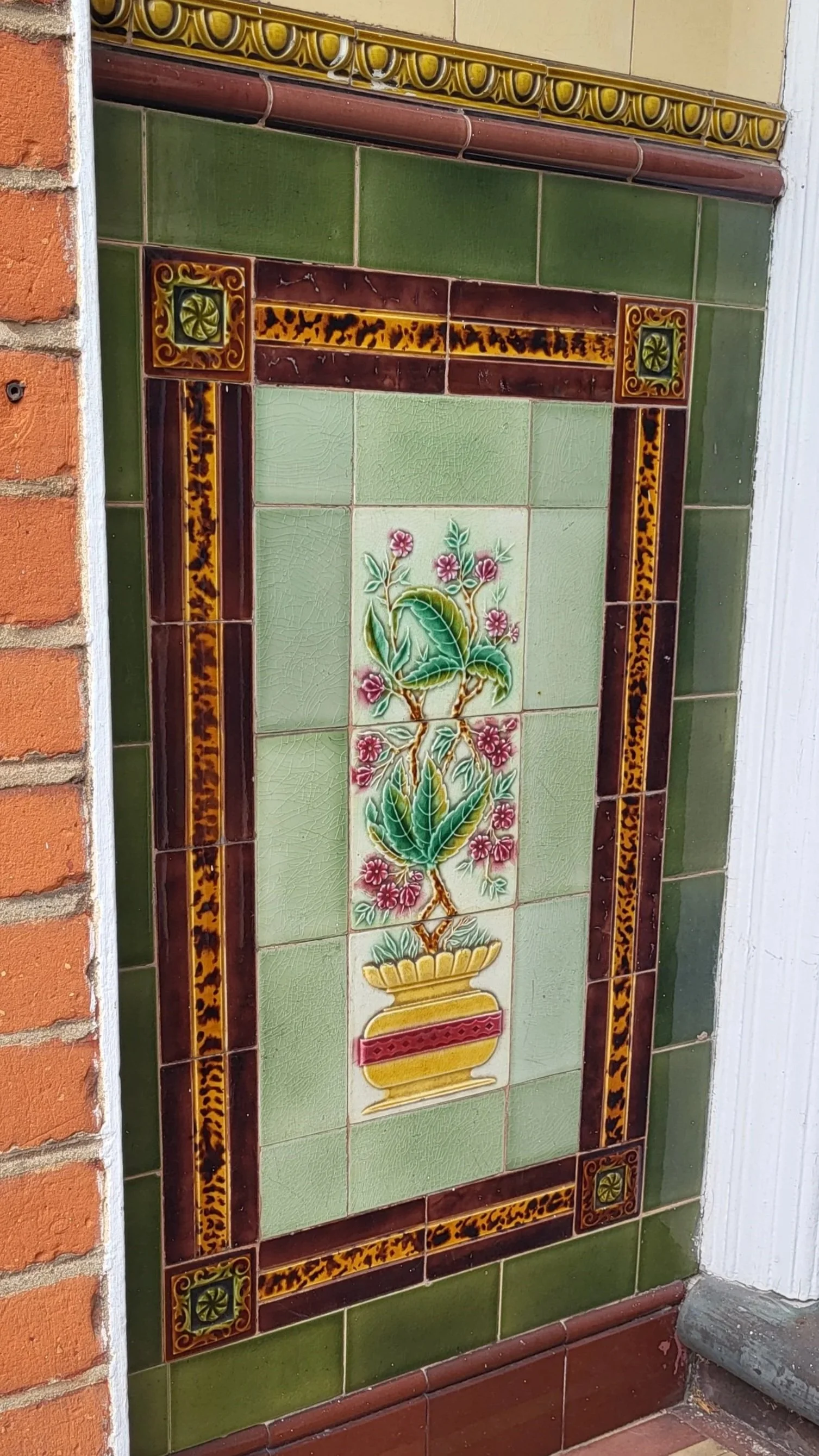 Decorative tiled wall with a potted plant design in the center, bordered by green, brown, and yellow tiles with intricate patterns.