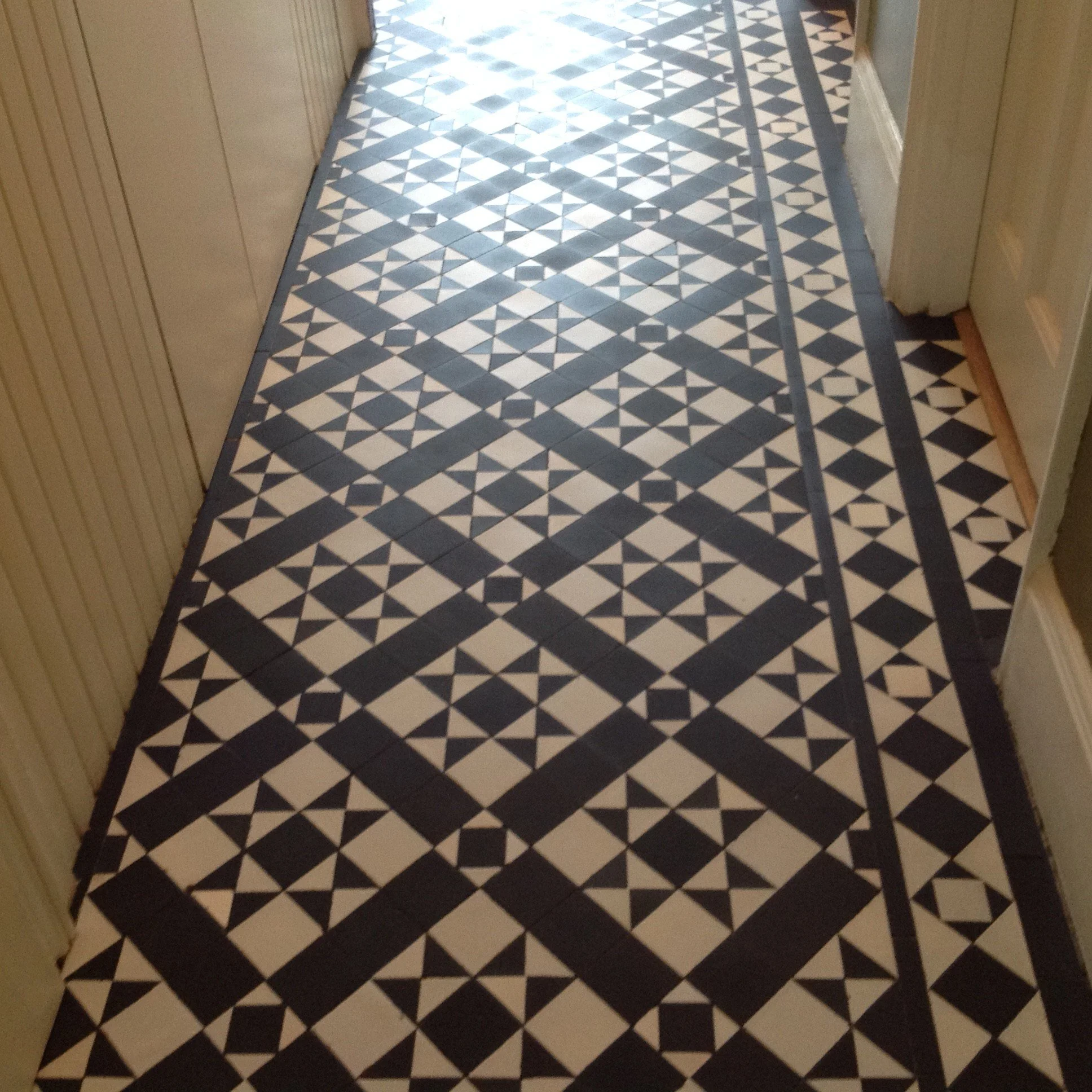 A hallway with patterned black and white floor tiles, featuring geometric and star-shaped designs, bordered by a narrow black tile edge.