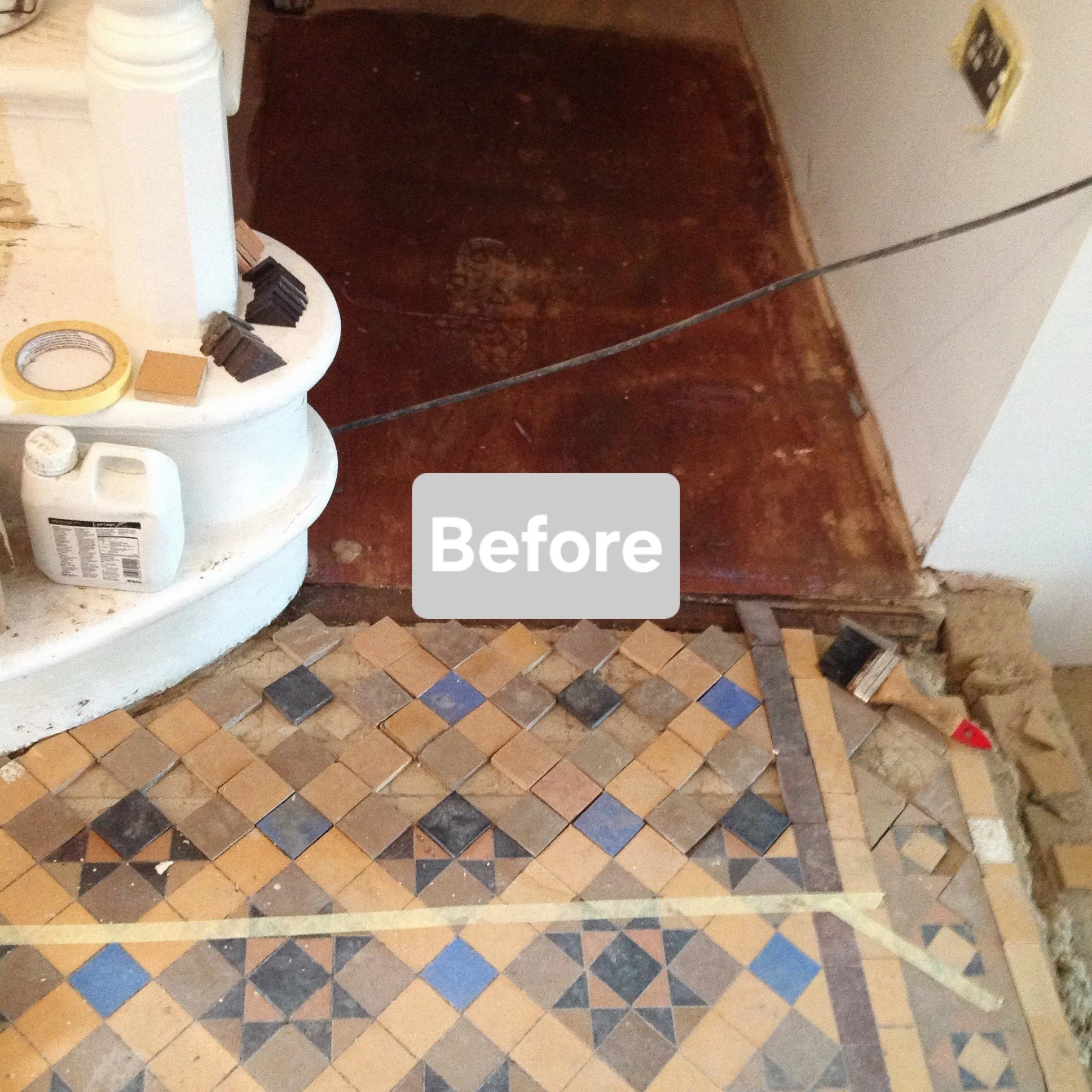 Floor with small decorative tiles, partially removed, in front of a wooden door with a metal strip, next to a white shelf with construction supplies, indicating a renovation in progress.