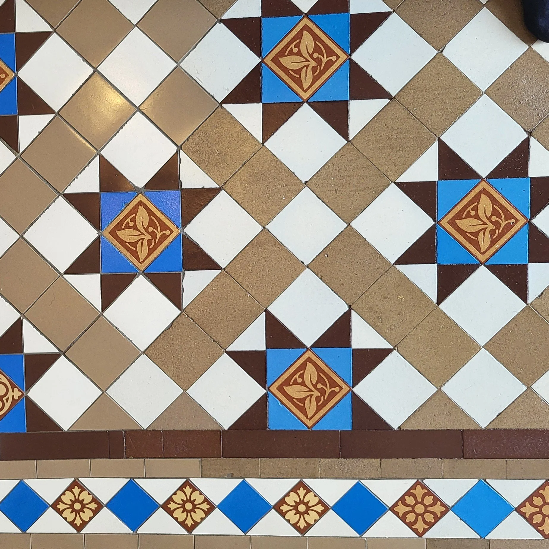 Patterned tile floor with geometric design, including diamond shapes in white, tan, blue, and brown, with floral motifs in the center of some blue squares.