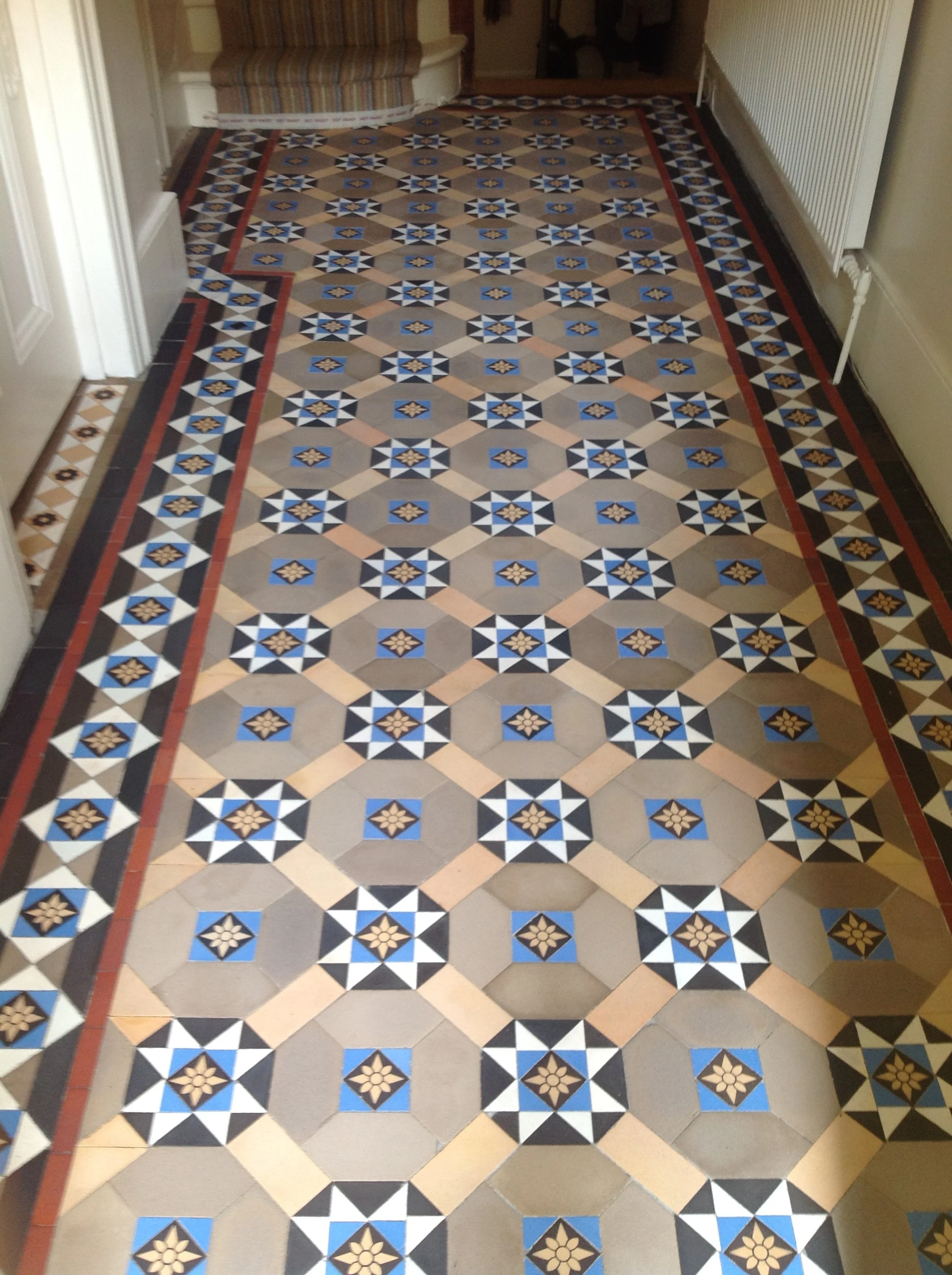 Decorative patterned tile floor in a hallway with geometric designs, featuring shades of cream, black, blue, and beige, bordered by a darker trim.