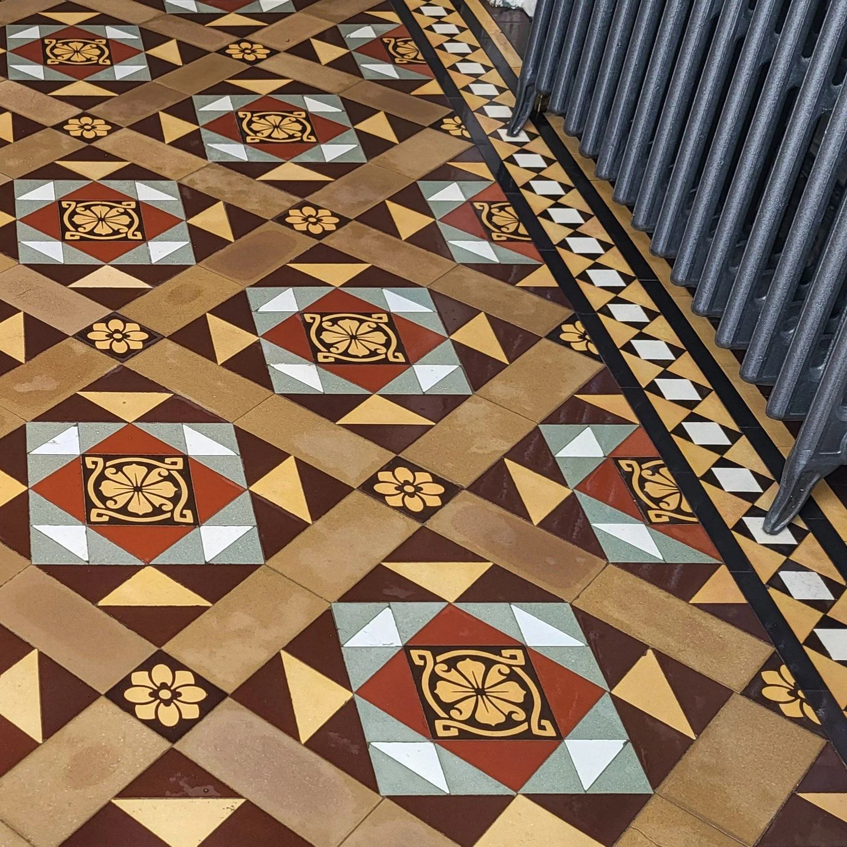 Decorative tile floor with geometric patterns and floral motifs in shades of brown, beige, black, white, and gray, with a metal barrier on the right side.