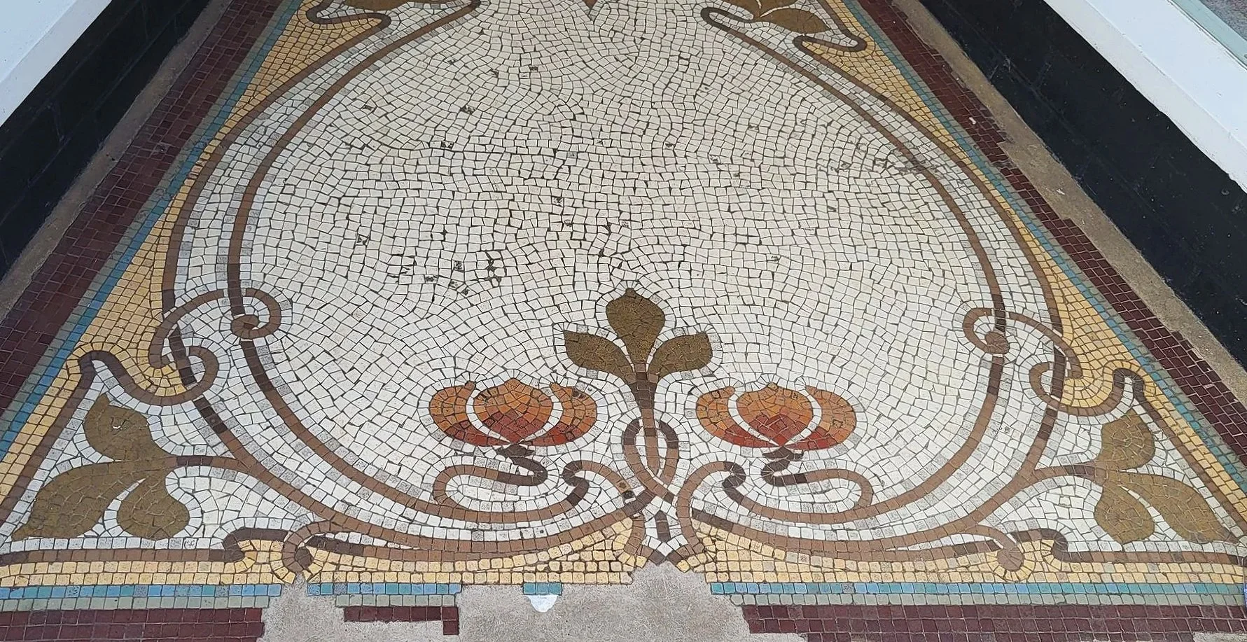 Decorative mosaic tile floor with floral and swirl patterns in earth tones, enclosed by a black and white border, some tiles are cracked or missing.