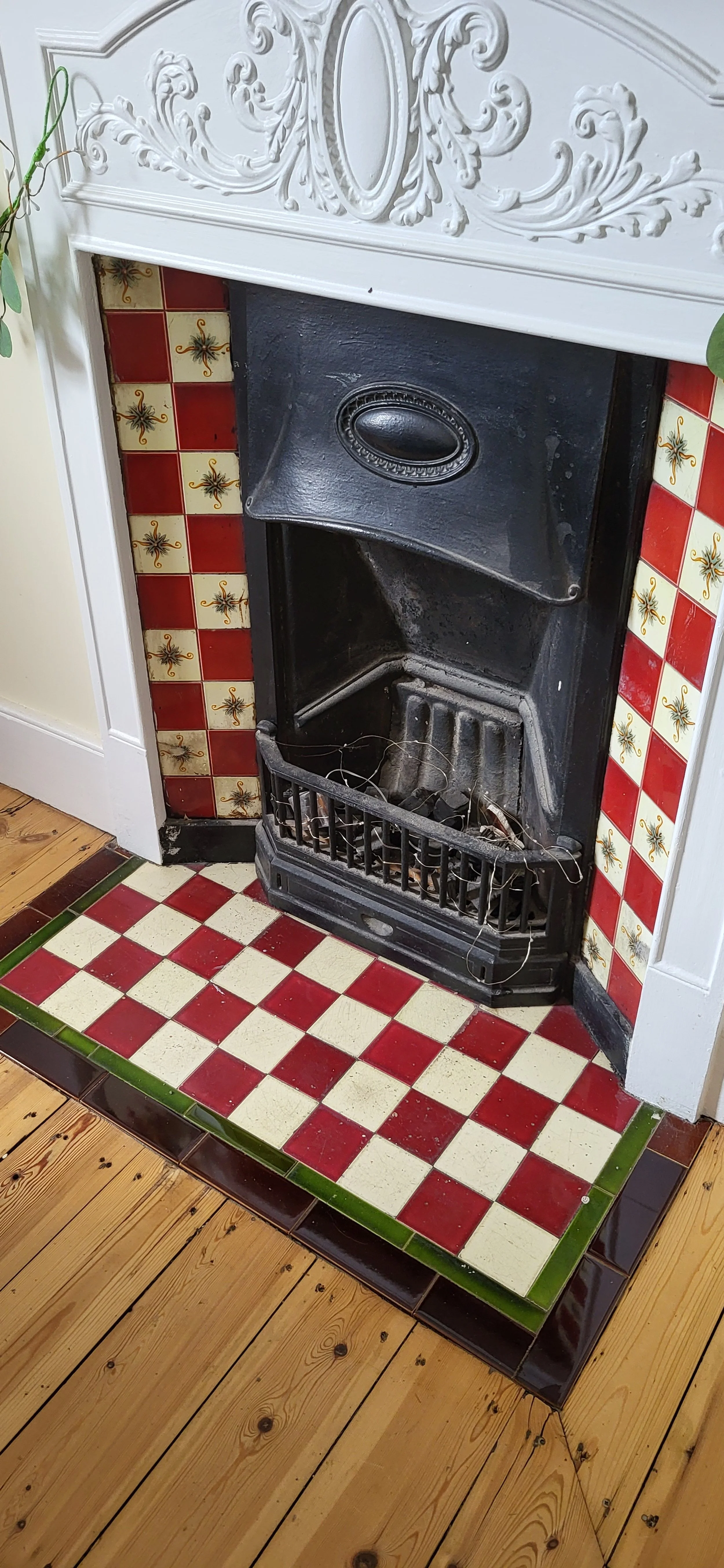 A fireplace with a black cast iron insert surrounded by red and yellow decorative tiles, a wood floor, and trim with plant vines growing nearby.