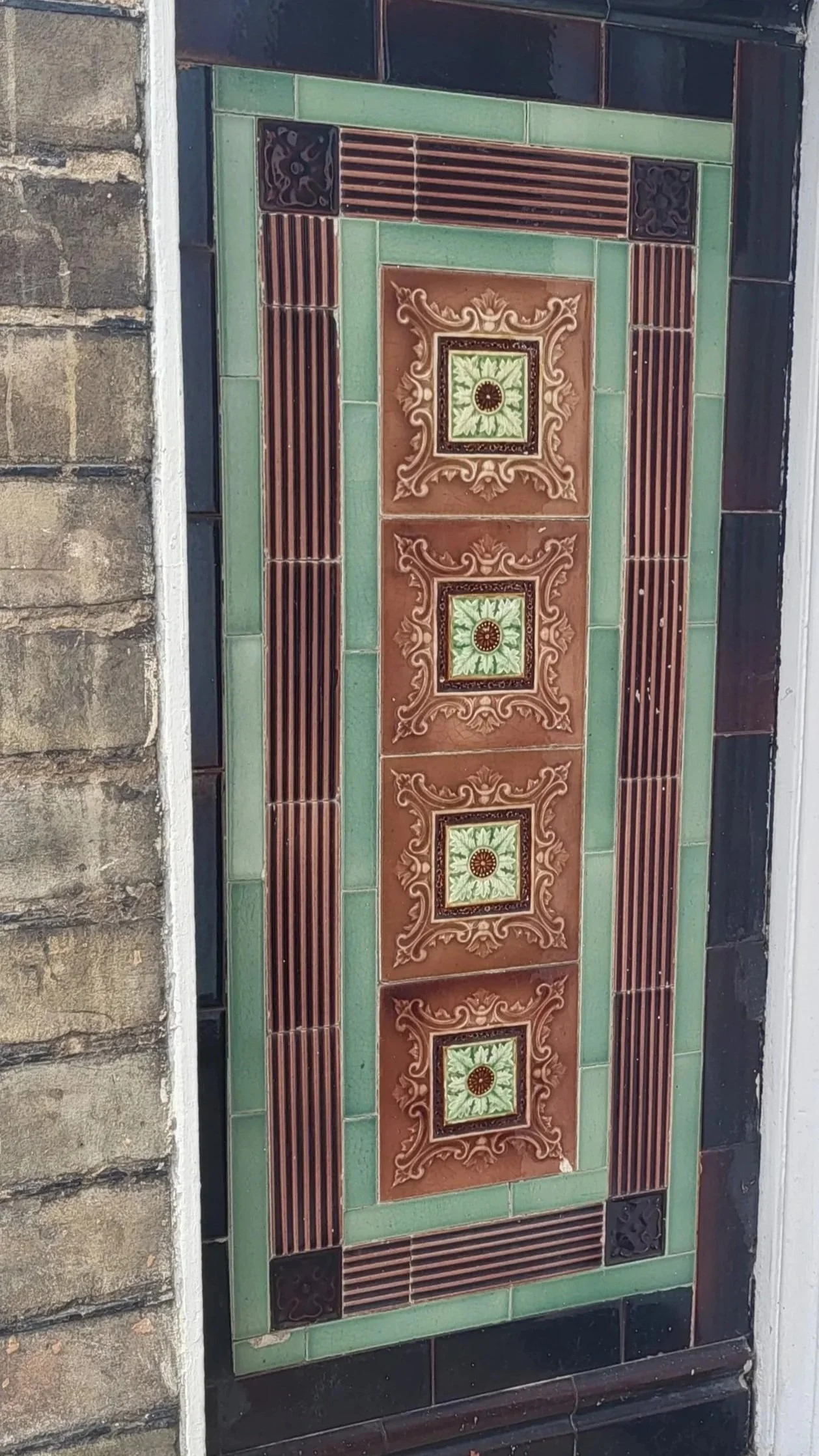 Decorative ceramic tile inlay on a wall with geometric and floral patterns in green, brown, and black colors.