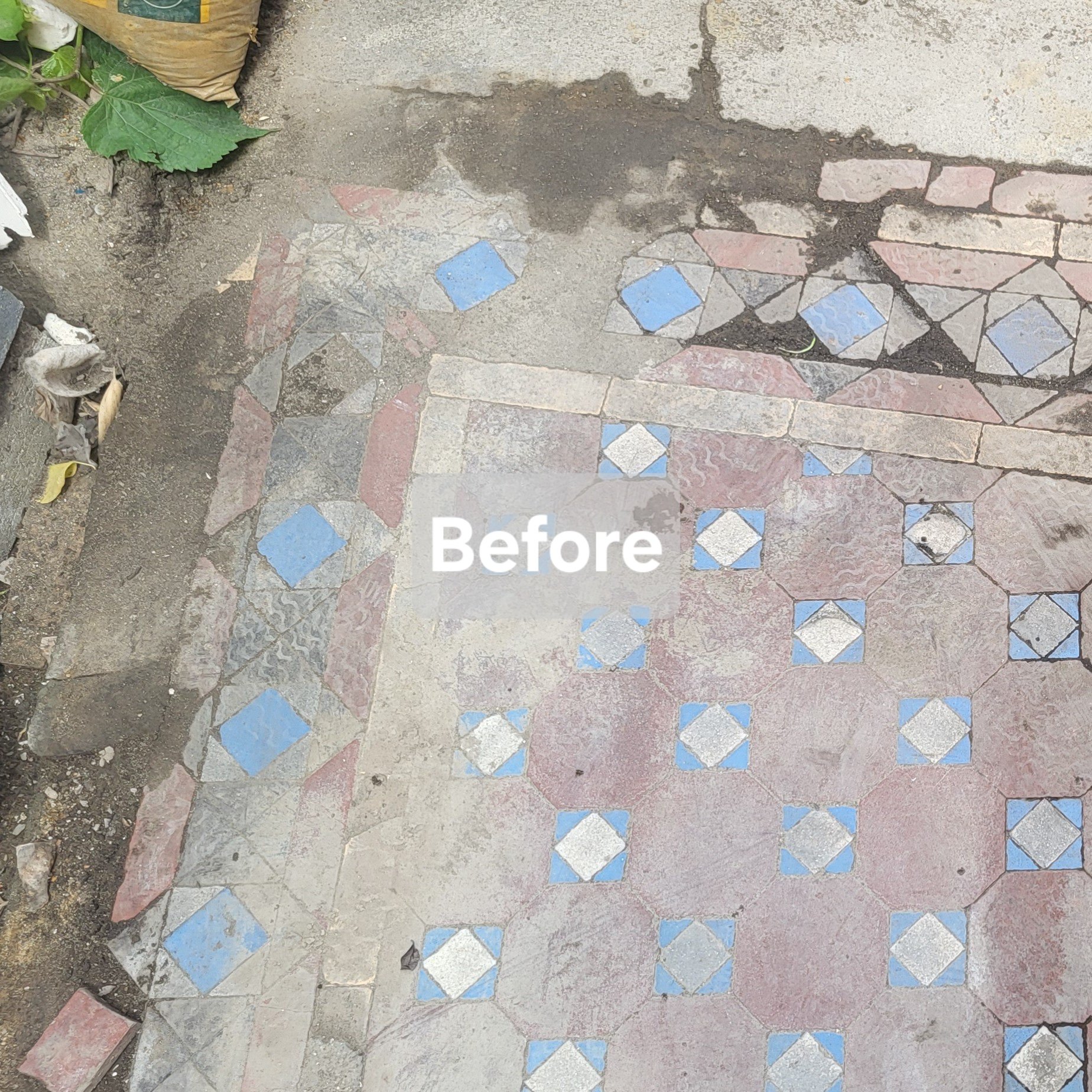 A section of an outdoor decorative tiled sidewalk with blue and red tiles, some of which are cracked or missing, and dirt and debris around the edges.