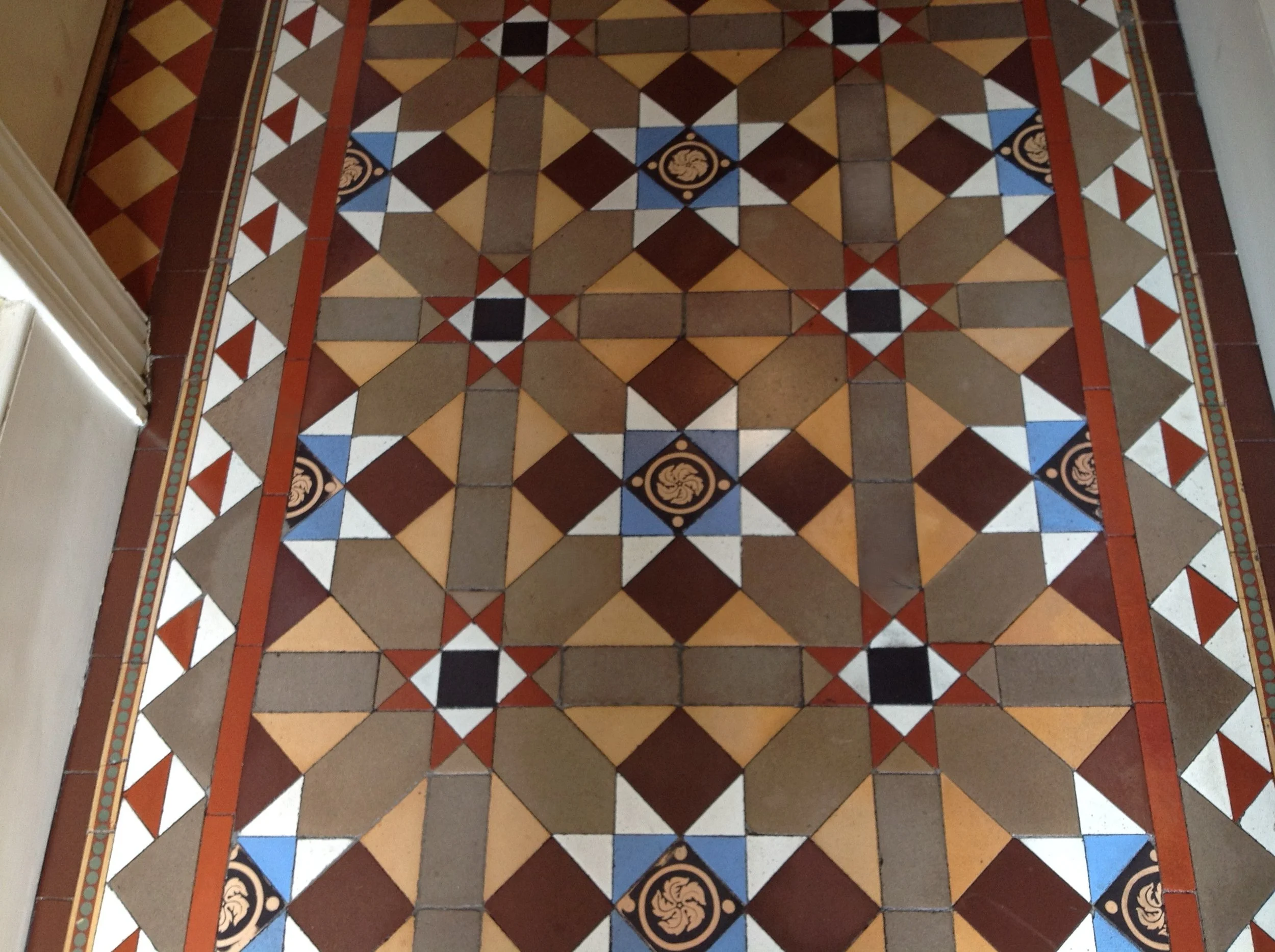 Colorful geometric patterned tile floor with a border design, featuring squares, triangles, and decorative motifs in shades of brown, blue, white, black, and reddish-orange.