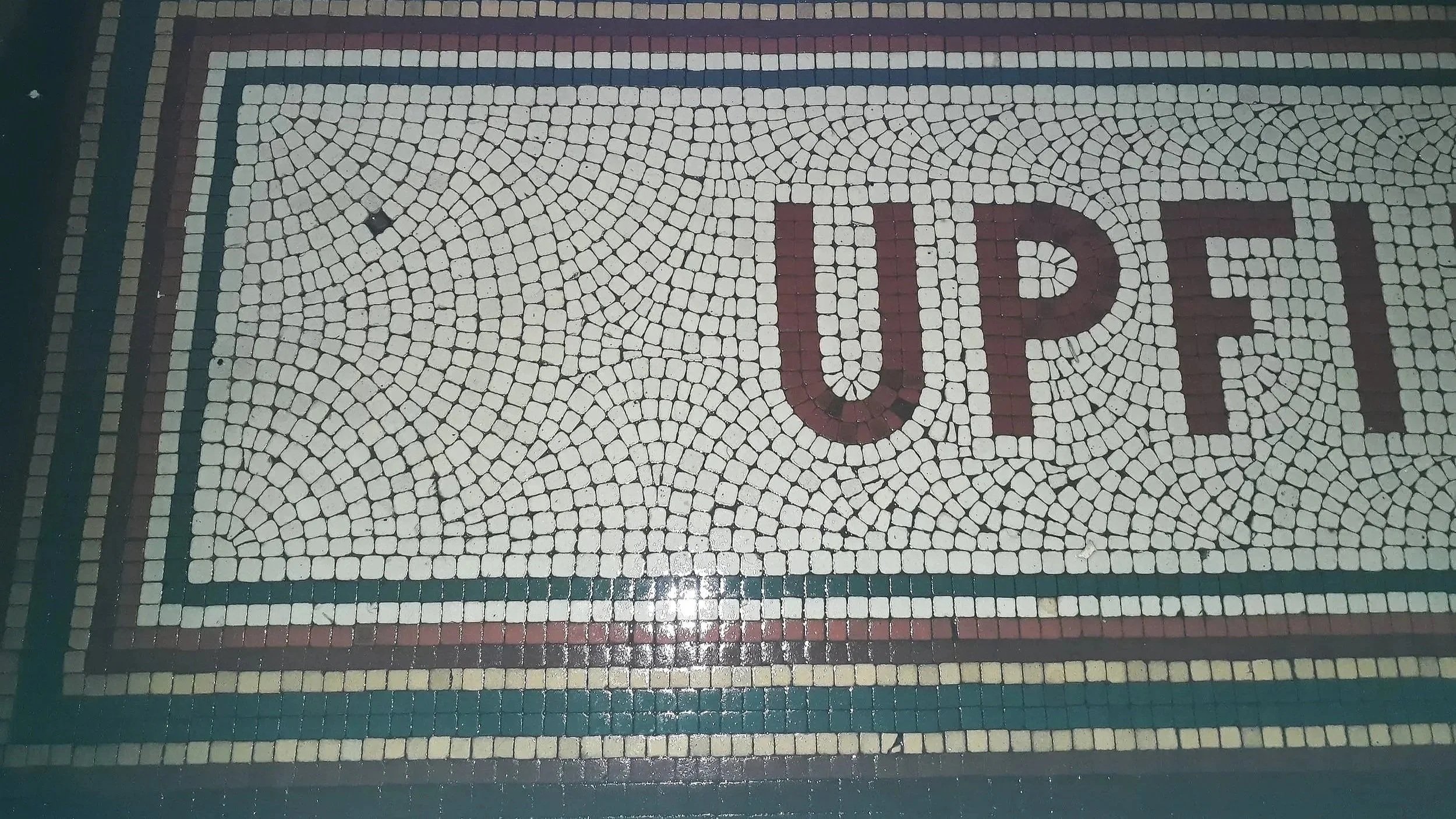 Mosaic floor with the word 'UPPER' spelled out in large dark red and purple tiles amidst smaller white tiles, bordered by concentric rectangular patterns in various colors.