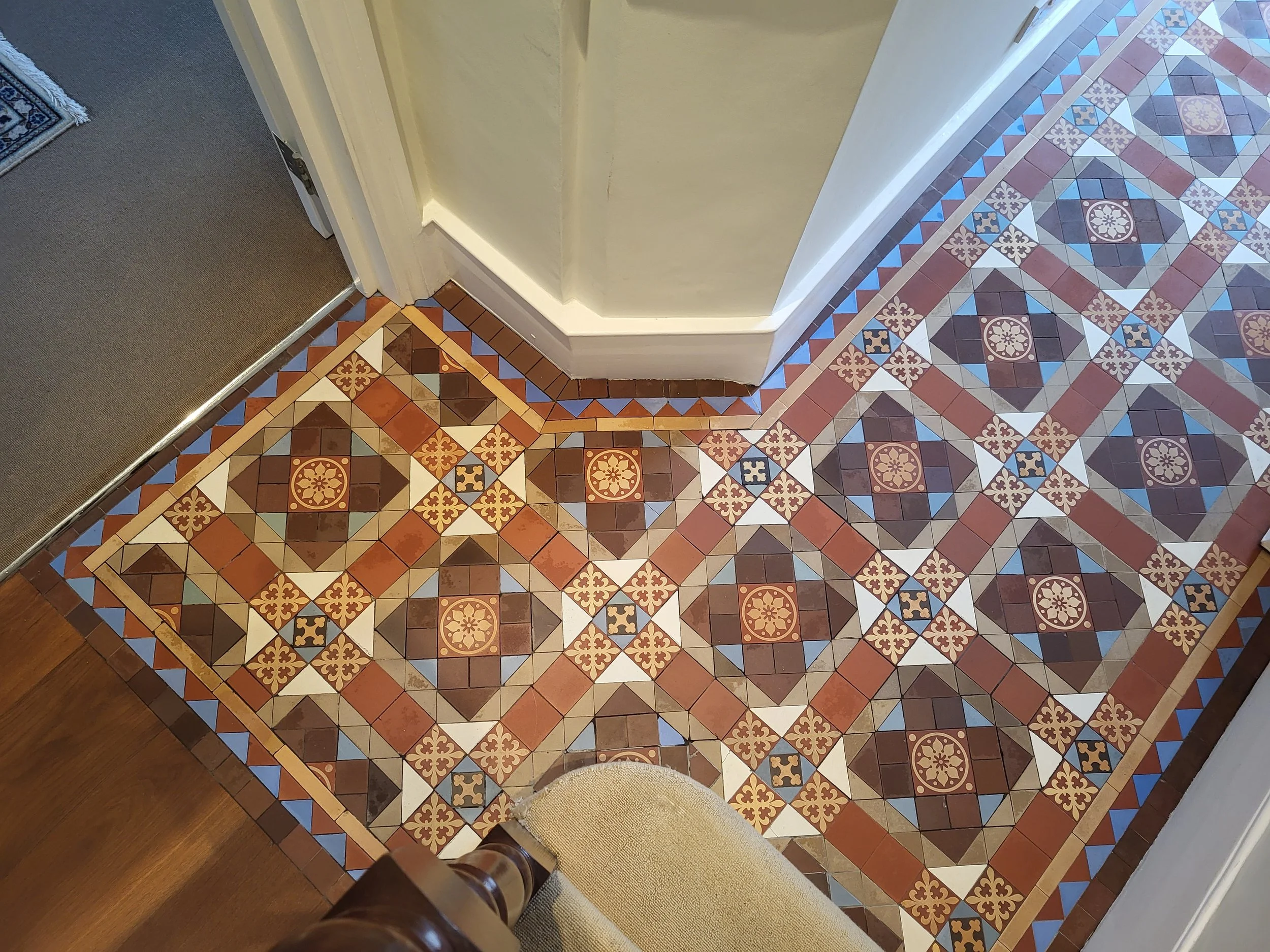 Decorative patterned tile flooring in a hallway with a corner and a portion of a cushioned chair visible.