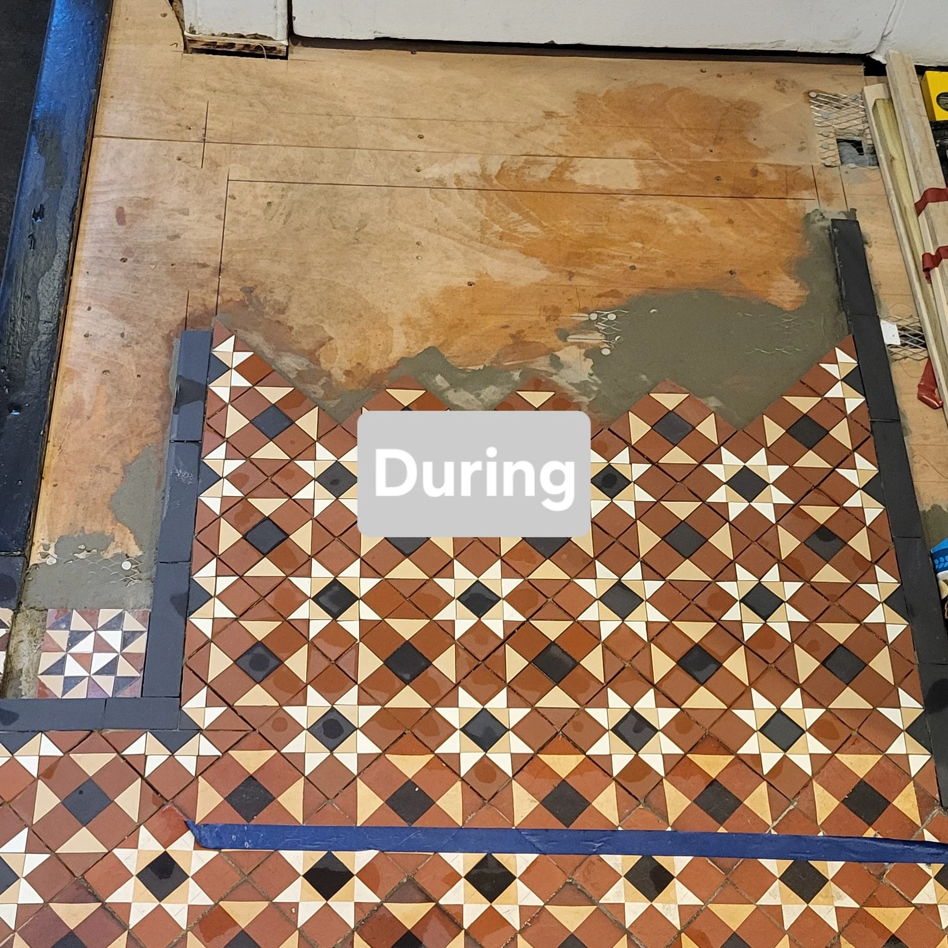 Tile flooring in progress with a geometric pattern of brown, white, black, and beige tiles, partially installed on a wooden subfloor during construction.