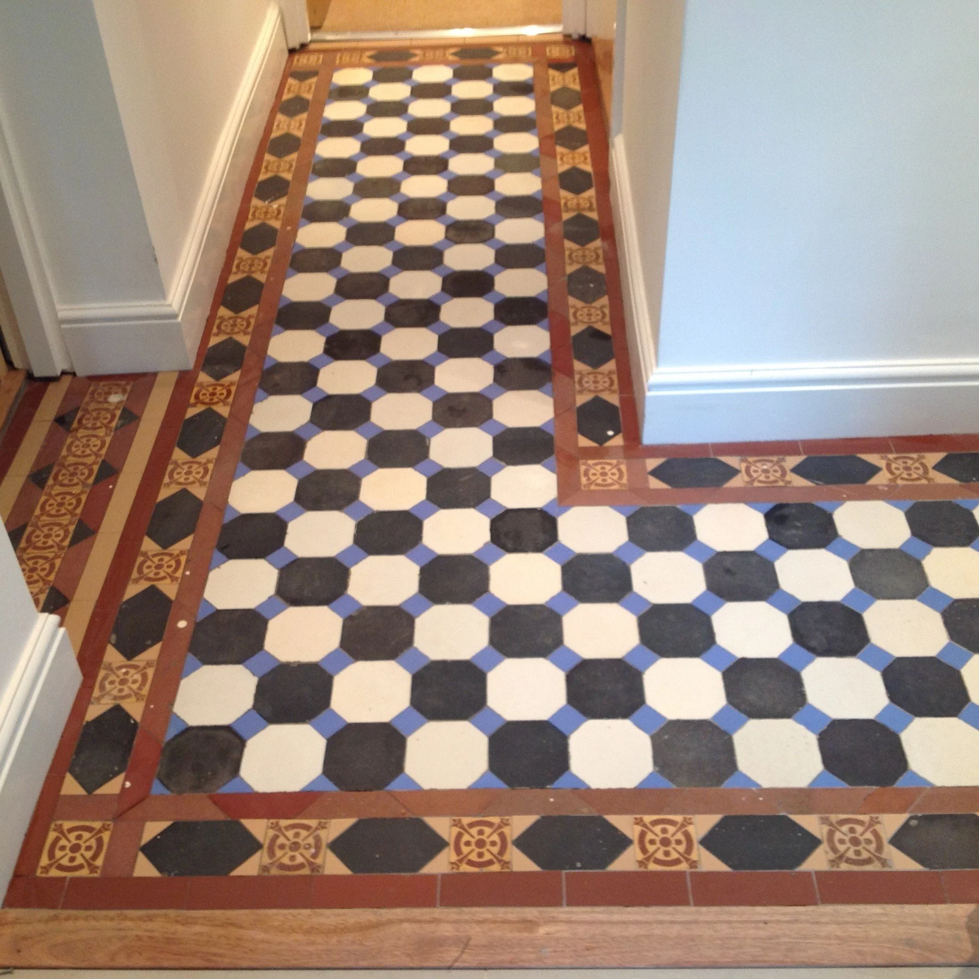 Patterned tile flooring in a hallway with black, white, and blue tiles, framed by decorative border tiles in red and yellow, along white walls.