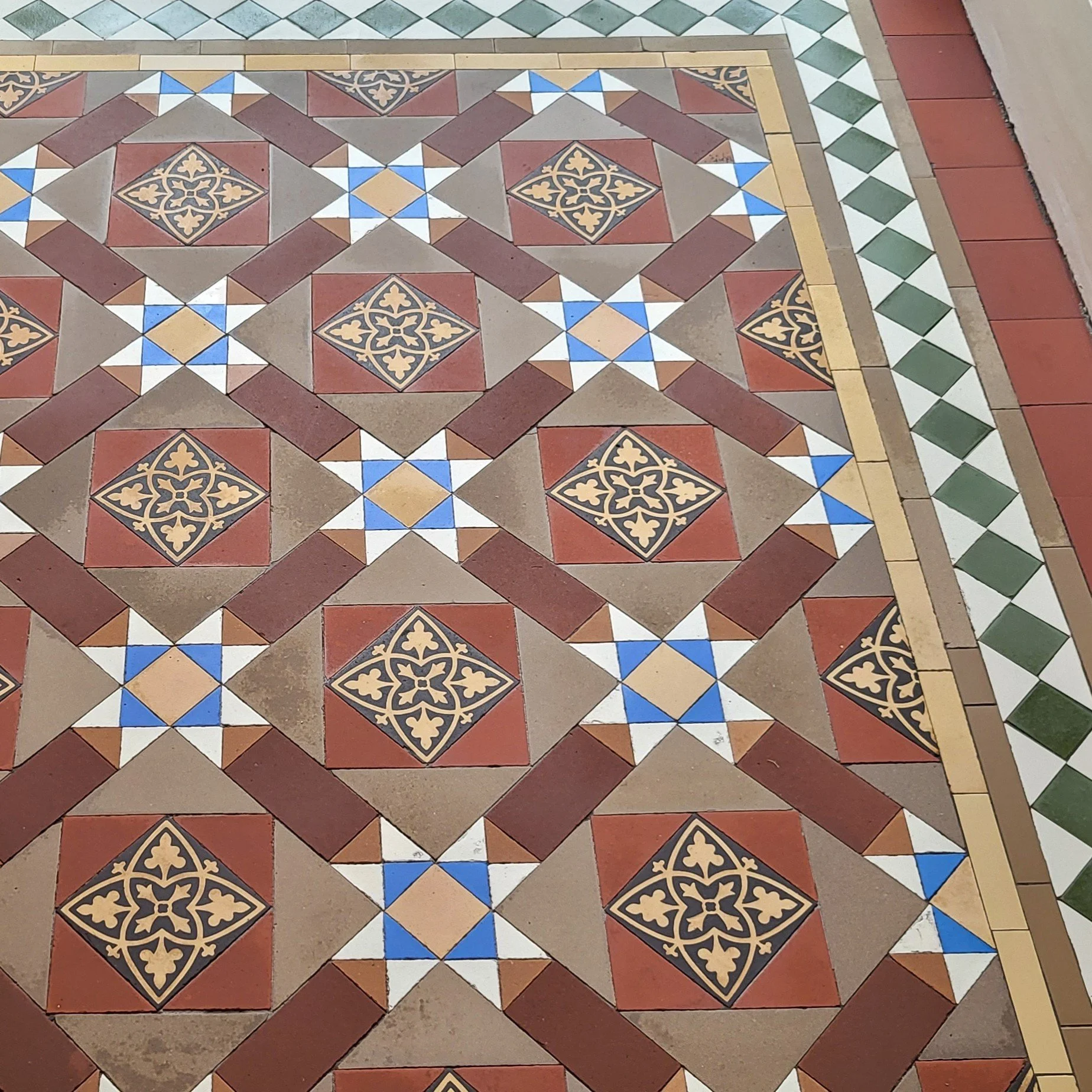 Decorative tiled floor with geometric patterns in red, beige, white, blue, black, and green. The main pattern features black tiles with yellow floral designs inside red squares, bordered by various smaller geometric shapes and patterns.