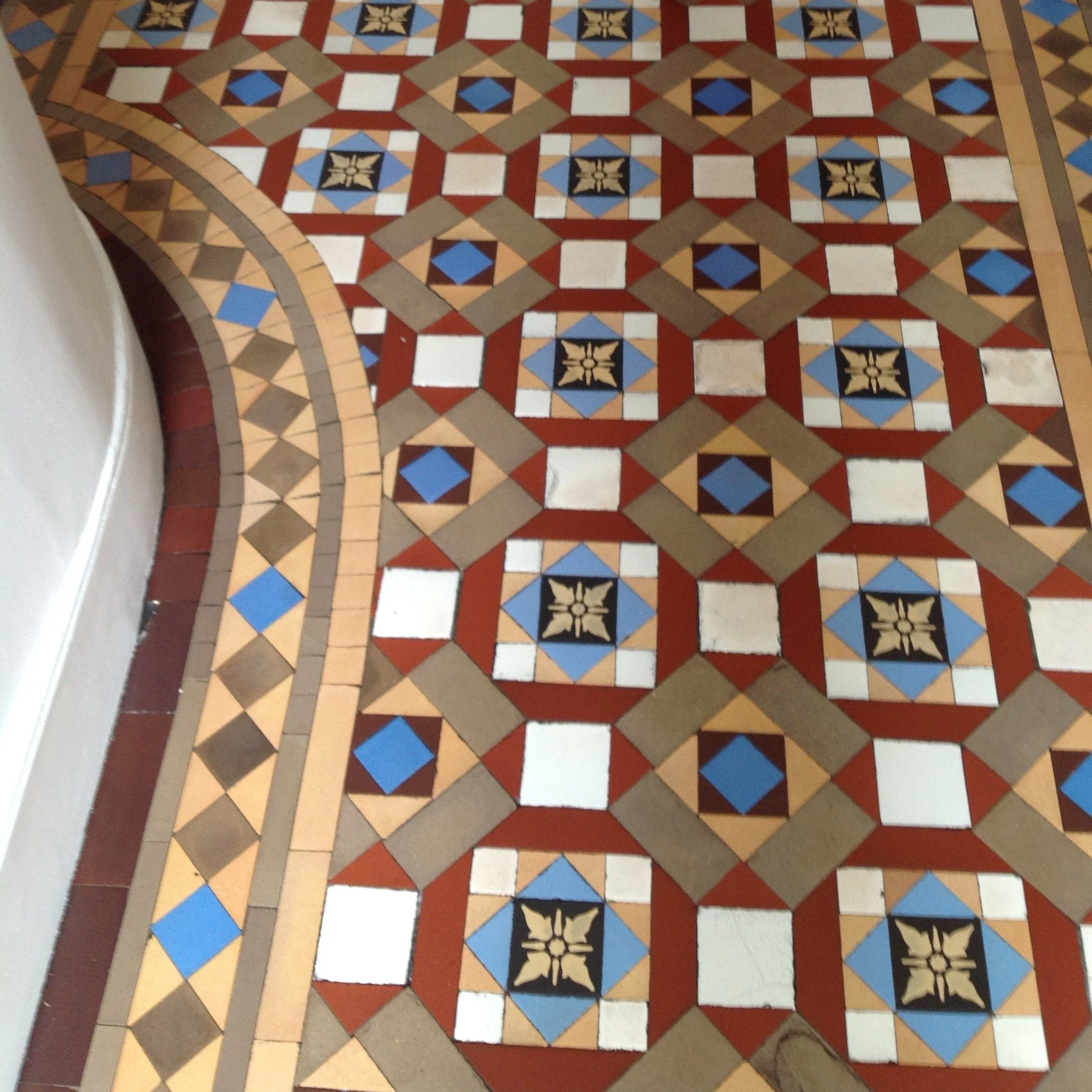 Colorful decorative tile floor with geometric pattern in beige, red, black, blue, and white, curving edge near a white wall.