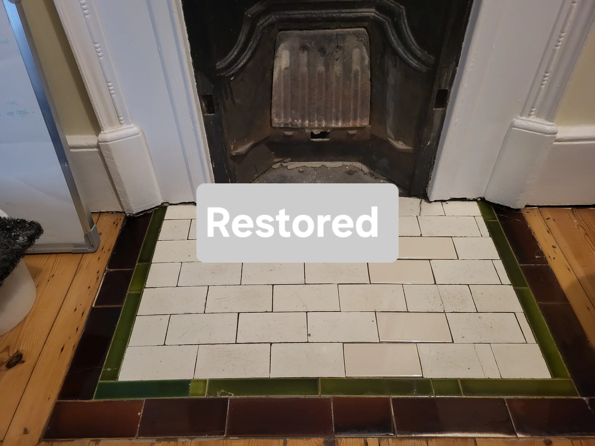 A fireplace with a blackened metal interior, surrounded by a white painted brick mantel, and a white tiled hearth with a green border, with the word "Restored" overlayed in the center.