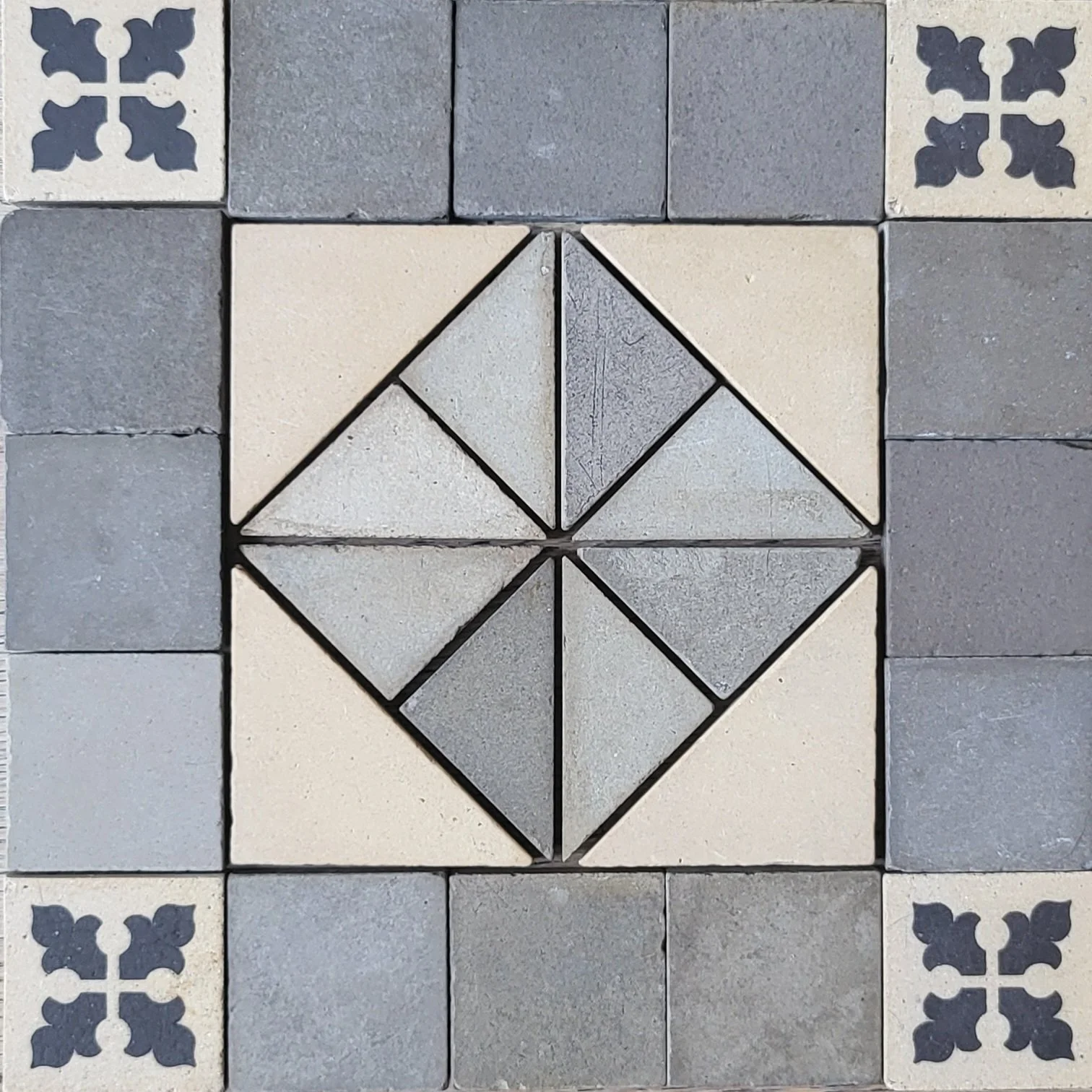 Patterned tiled floor with square and diamond shapes in shades of gray and cream, with decorative corner tiles featuring black floral motifs.