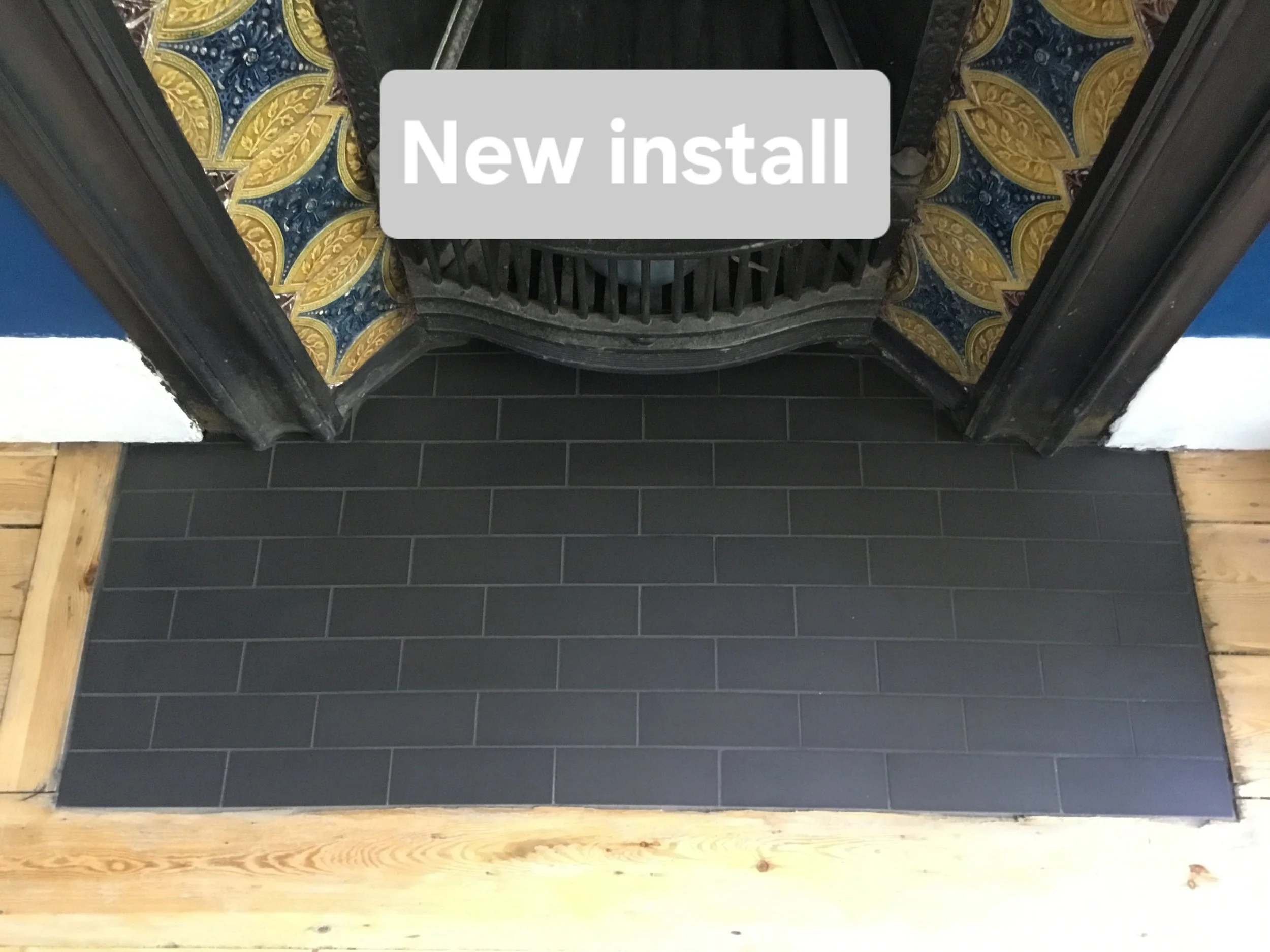 Newly installed dark gray tile hearth in front of ornate black fireplace with colorful decorative tiles and a fire grate, with wooden flooring around the hearth.