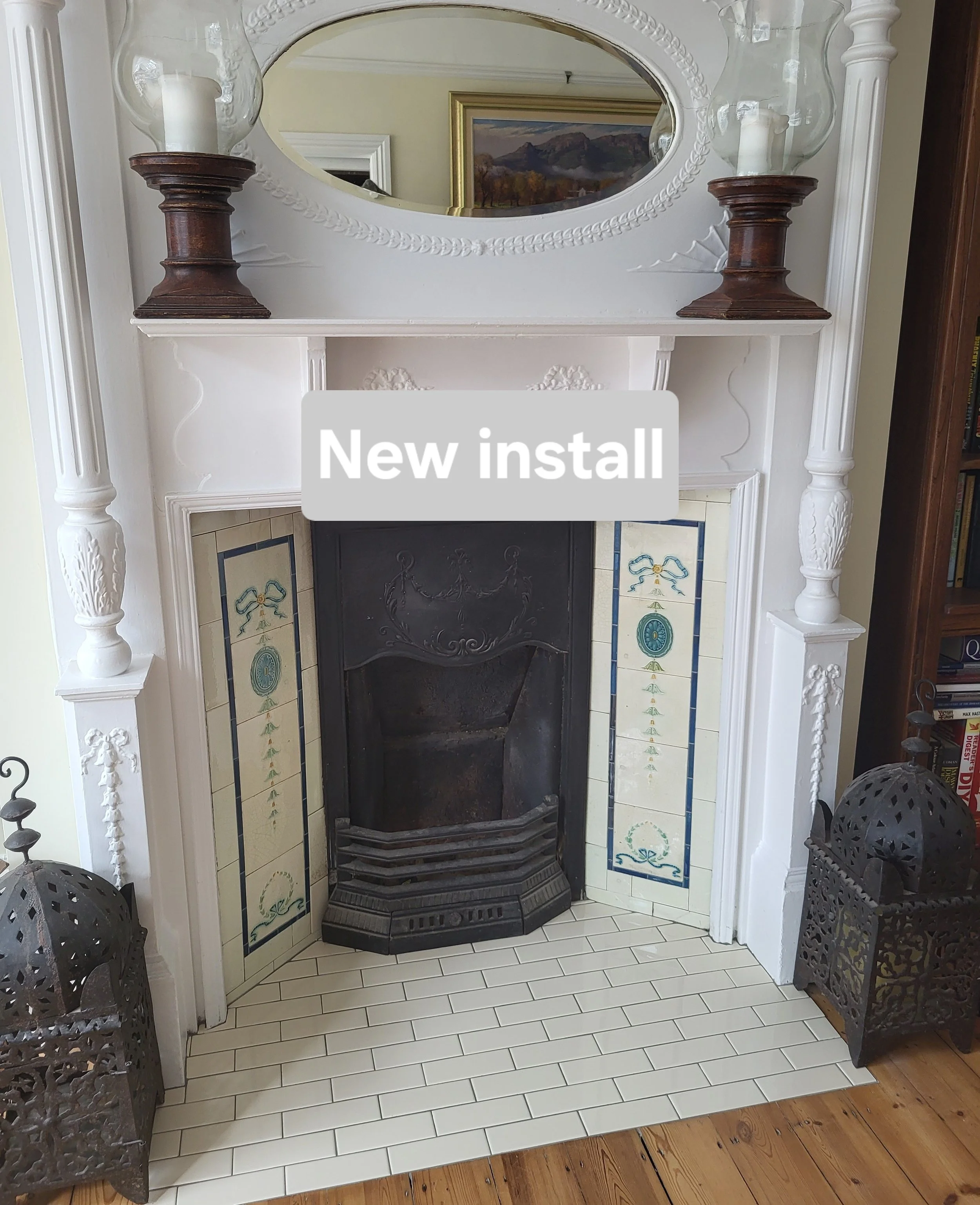 A fireplace with a decorative white wooden mantel, an oval mirror above it, and two large glass hurricane lamps on wooden stands on either side. The fireplace opening has an ornate metal fireguard and the surround is adorned with decorative blue and 