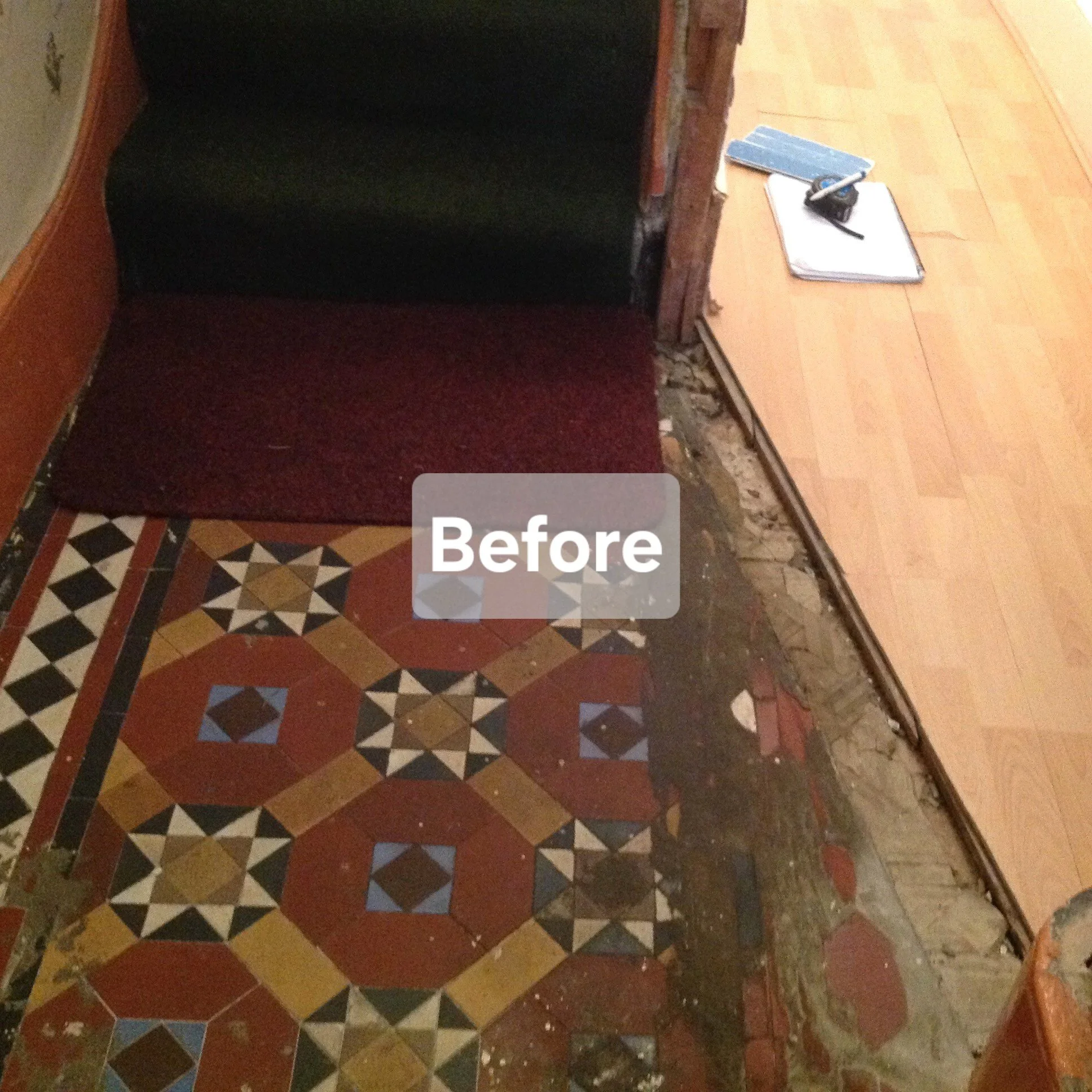 A partially renovated entryway with a new wooden floor on the right side, an old patterned ceramic tile floor on the left side, and a red doormat at the entrance. The transition between floors is rough, with visible construction and missing trim.