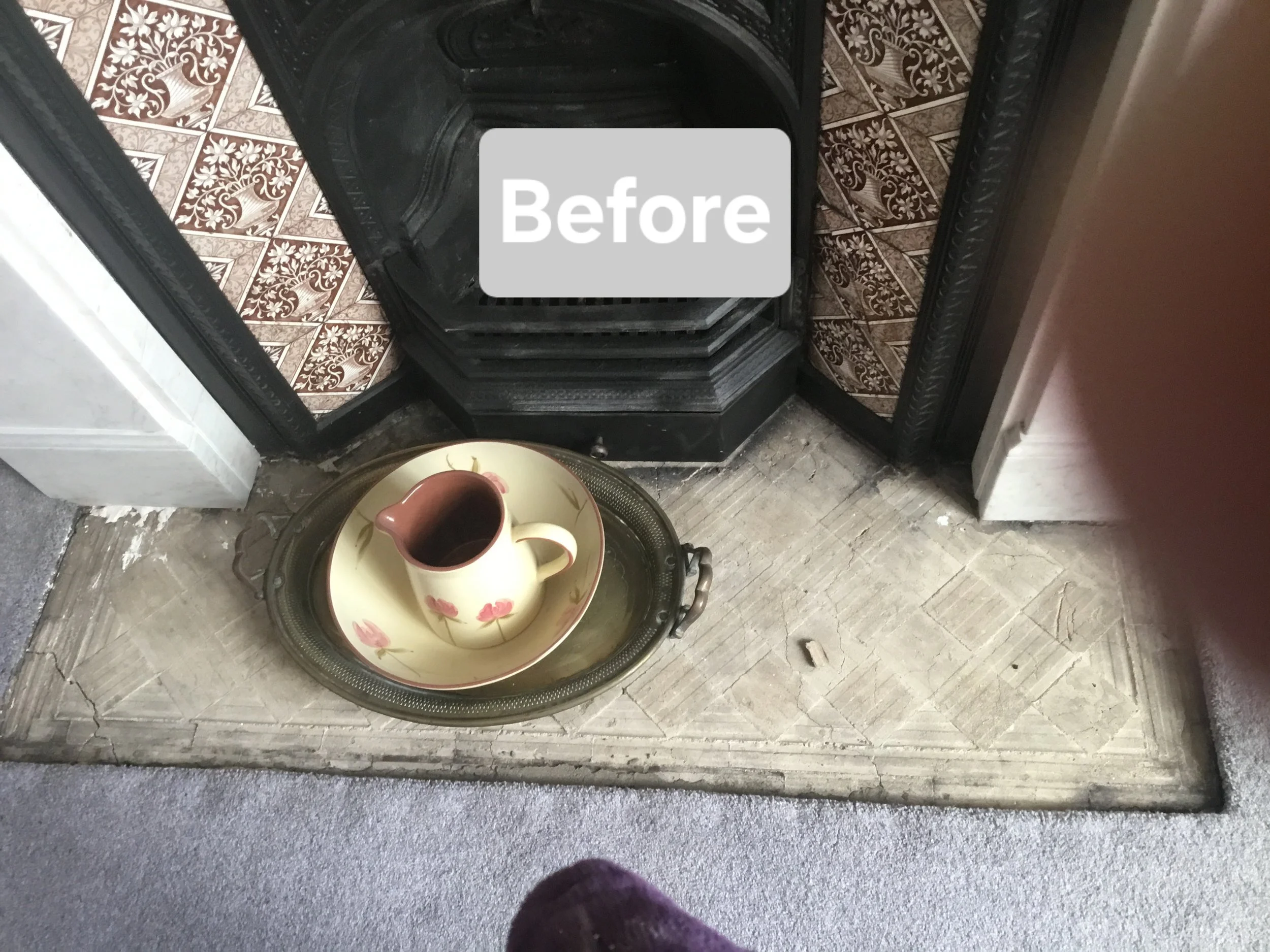 A fireplace with a black metal surround and a decorative border, with a rug or small mat in front of it. On the mat, there is a metal tray holding a ceramic pitcher and a matching bowl with a floral pattern.