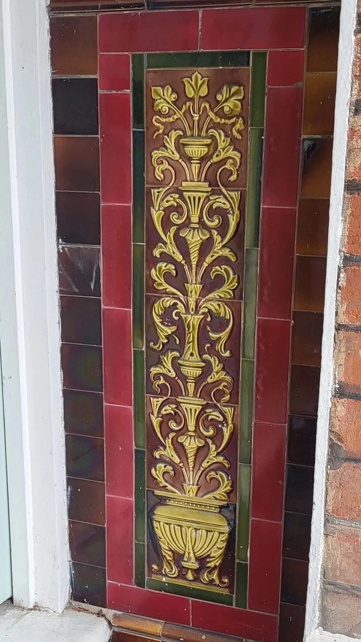 Decorative tile mural with a yellow and gold vase and swirling plant motifs on a red tiled background, bordered by green tiles.