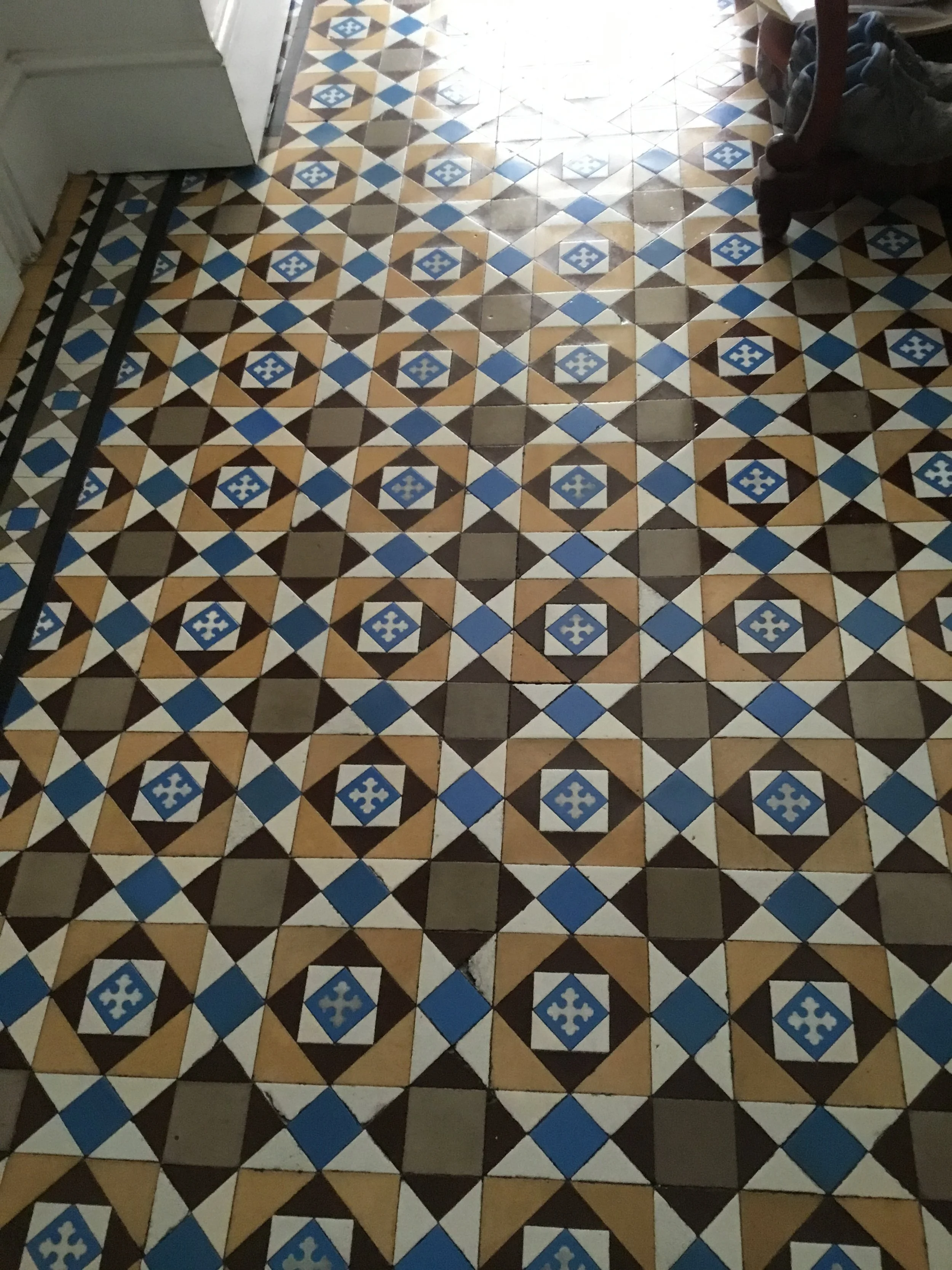 Patterned vintage ceramic tile floor with geometric designs in brown, blue, beige, black, and white, some tiles chipped or damaged.