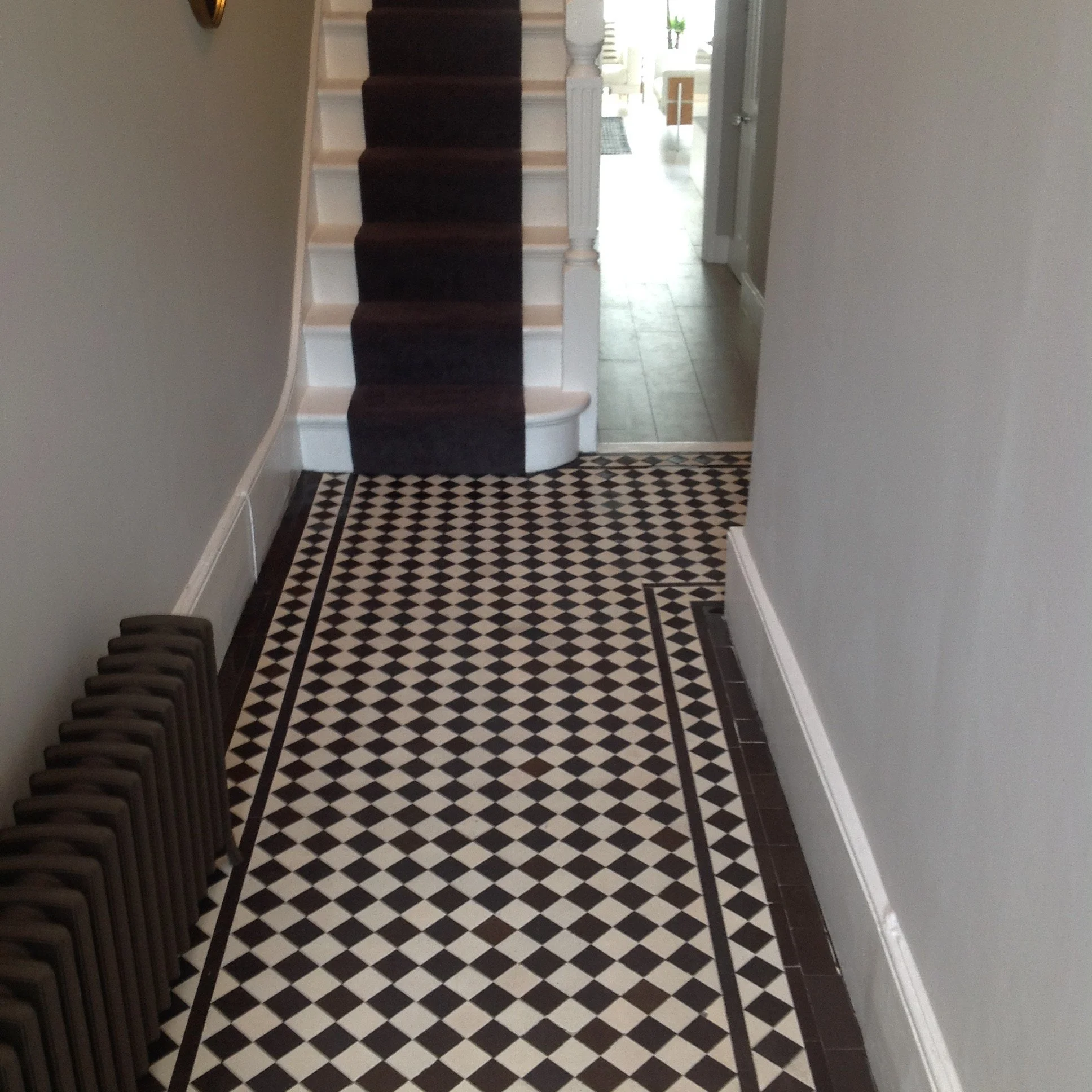 Entryway with patterned black and white tile floor, a radiator on the left, and stairs with a dark runner carpet leading upwards. There is a hallway visible in the background.