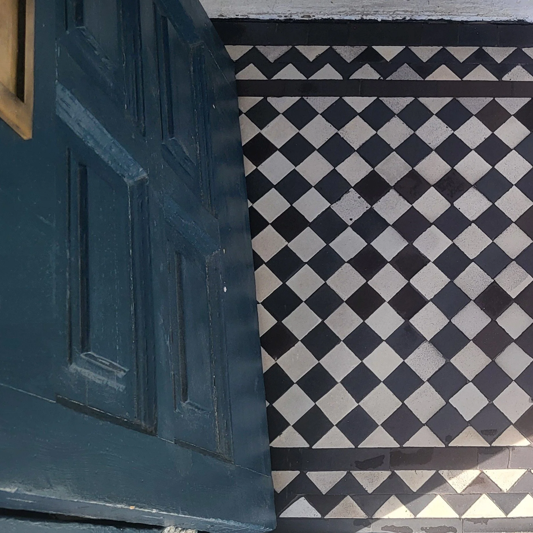 Close-up of a worn wooden blue door with a decorative panel, and a black and white checkered floor beneath it.
